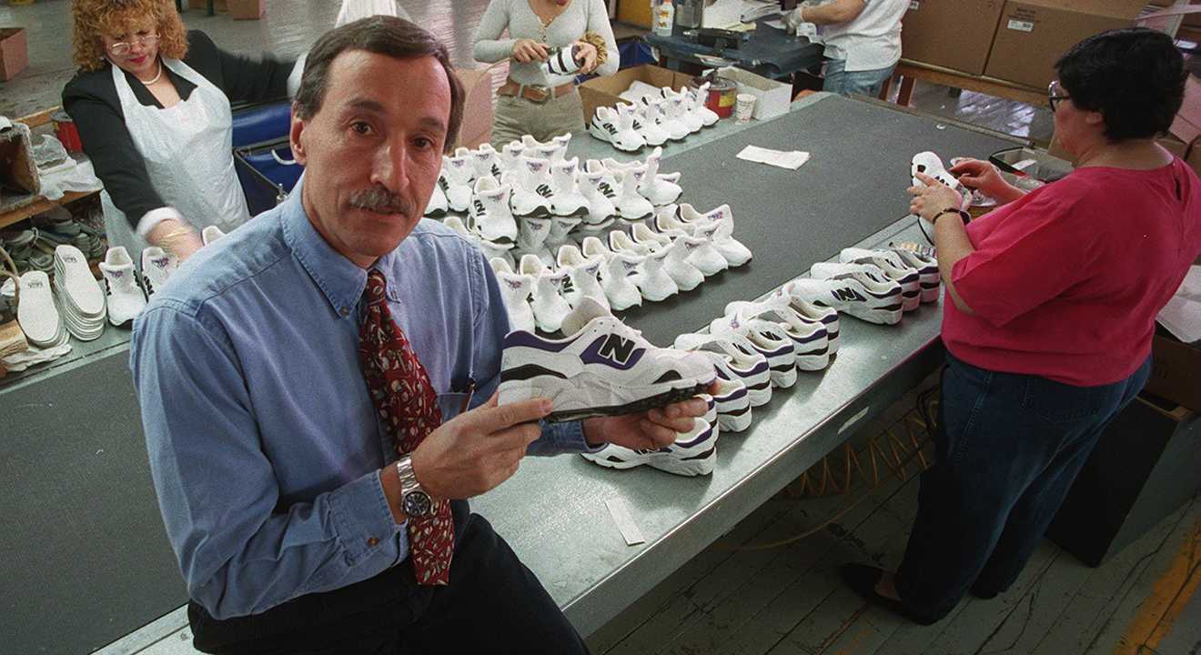 Mann in Werkstattszene hält New-Balance-Sneaker in die Kamera, im Hintergrund Reihen fertiger Schuhe