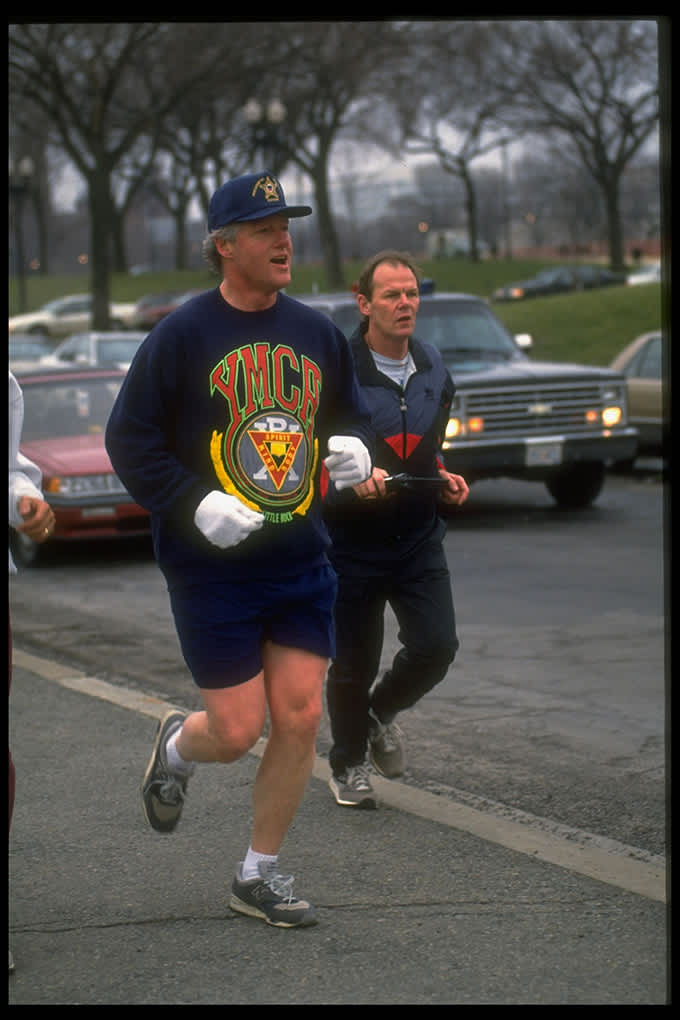 Bill Clinton im YMCA-Sweatshirt mit Trainingspartner auf Straße, umgeben von parkenden Autos