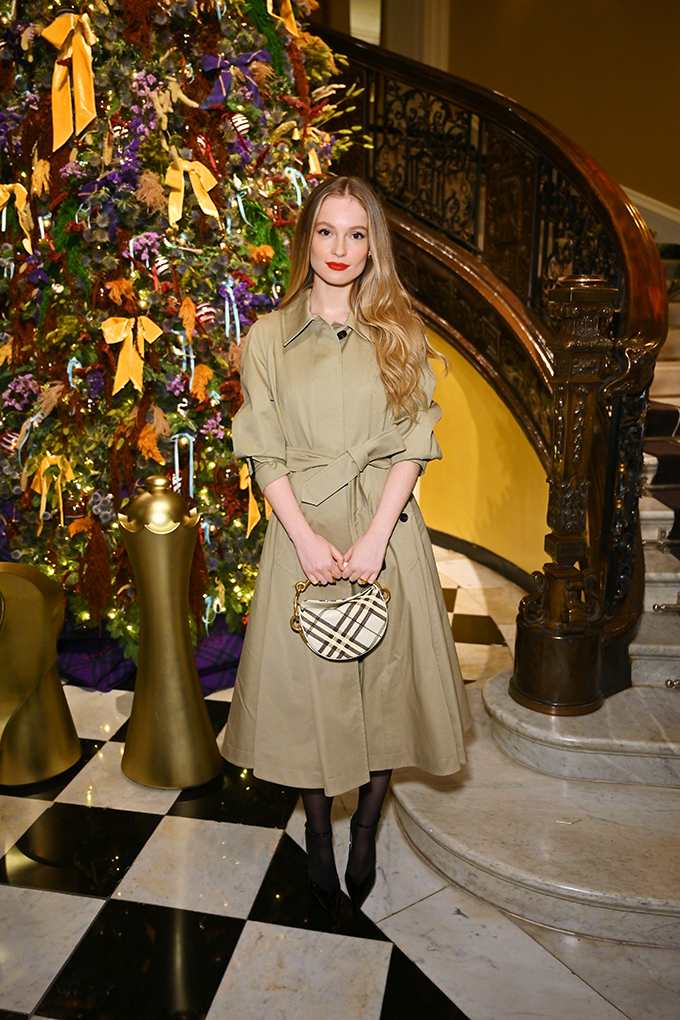 Schauspielerin Hannah Dodd trägt beiges Trenchkleid mit Karo-Tasche und schwarzen Stiefeletten vor Weihnachtsbaum