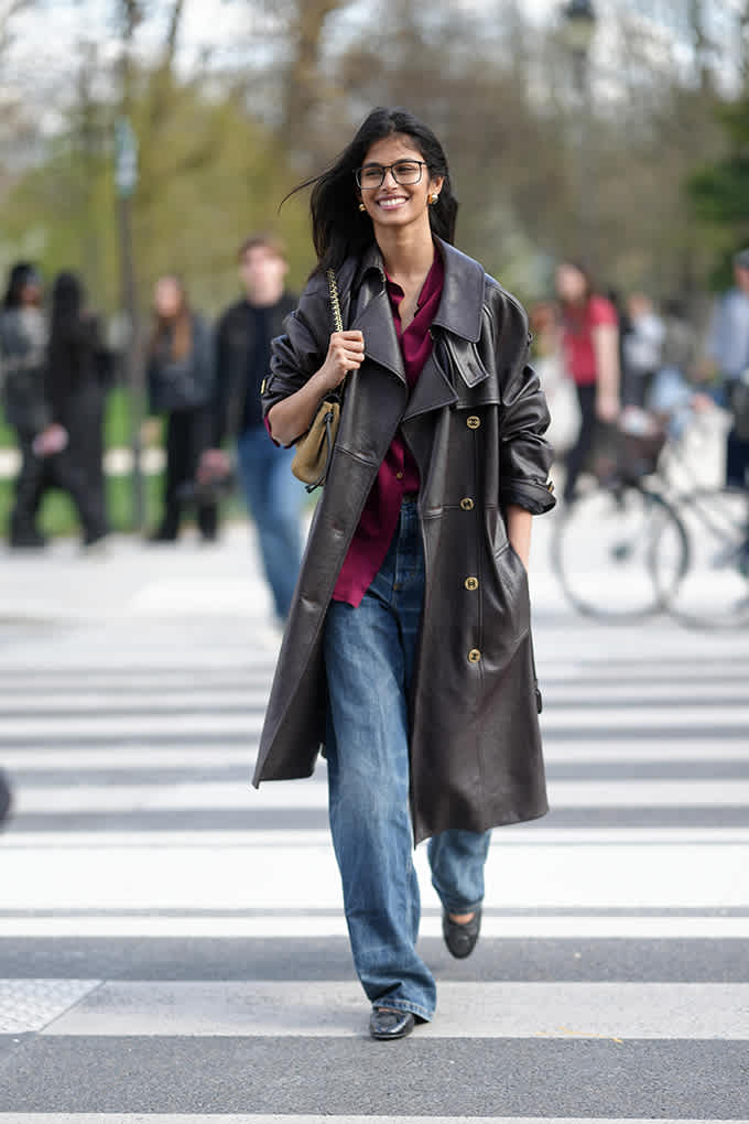 Bhavitha Mandava mit langem Ledertrenchcoat, weiter Jeans und Loafern auf Zebrastreifen