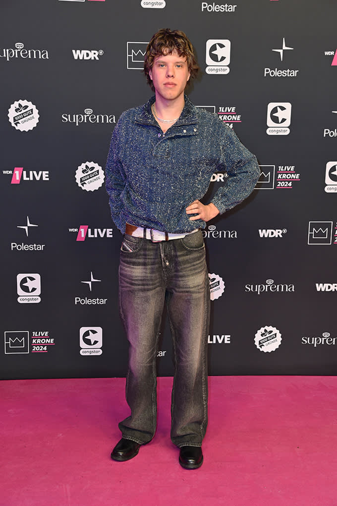 Rapper Zartmann in blauem Bouclé-Hemd, weiter Waschjeans und schwarzen Lederschuhen vor Fotowand