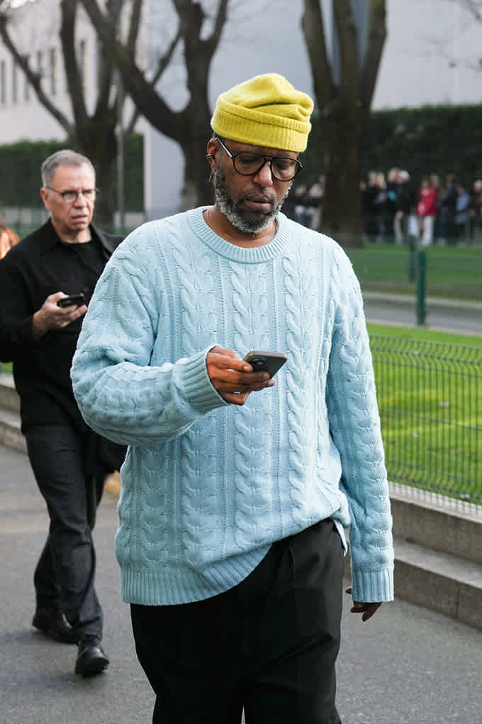 Mann mit gelber Mütze und Brille in hellblauem Strickpullover hält ein Smartphone in der Hand