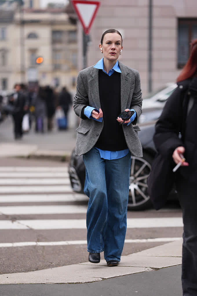 Eine Frau überquert eine Straße in grauem Blazer, schwarzem Strickpullover und weiter Jeans