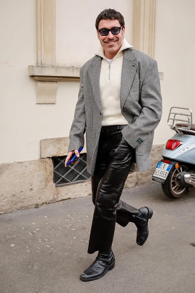 Ein Mann trägt einen grauen Blazer, weißen Strickpullover, schwarze Lederhose und Stiefel