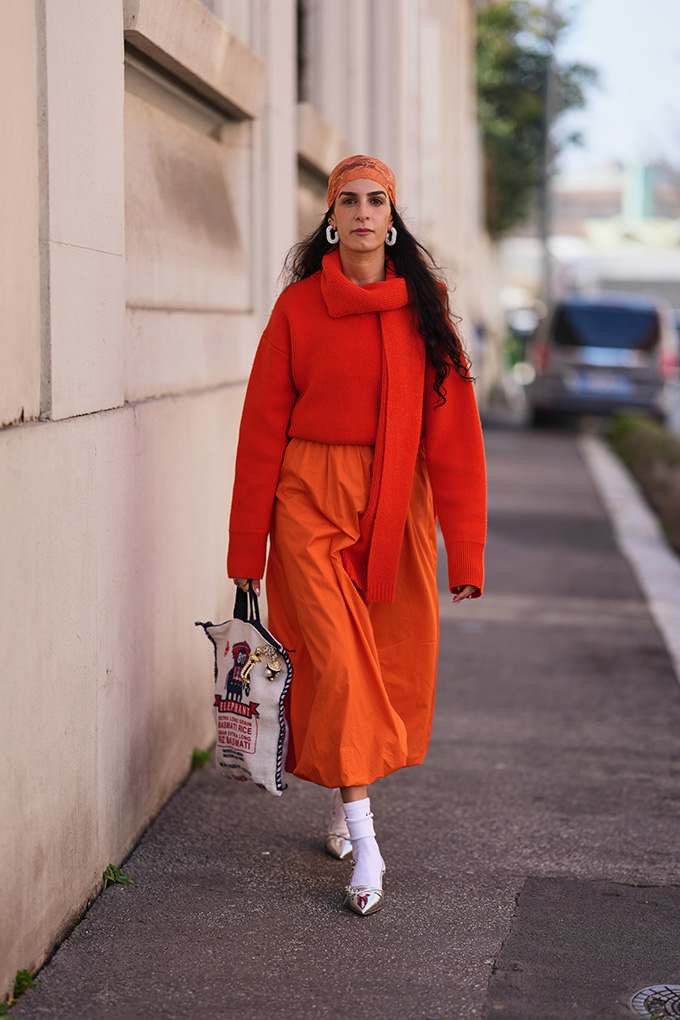 Frau mit orangefarbenem Kopftuch, rotem Pullover und orangem Rock läuft mit Tasche auf einer Straße