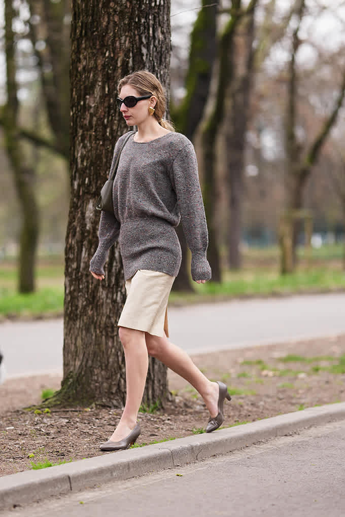 Frau mit grauem Pullover, hellem Rock und Sonnenbrille läuft an Bäumen entlang