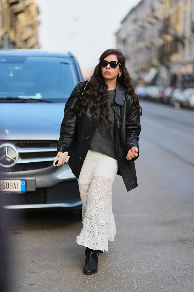 Eine Frau mit Sonnenbrille, schwarzer Lederjacke und langem Spitzenrock steht vor einem Auto