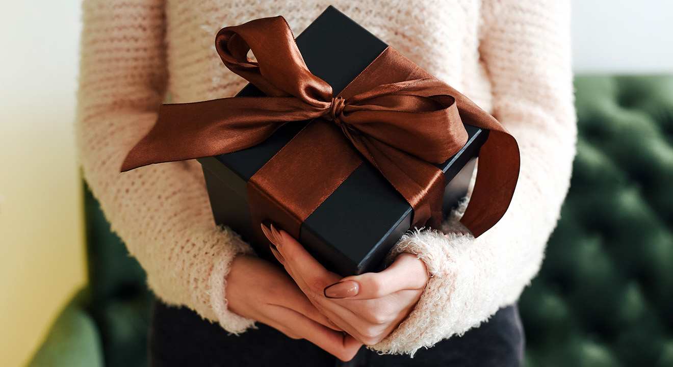 Hände im Flauschpullover halten schwarze Geschenkbox mit brauner Satinschleife