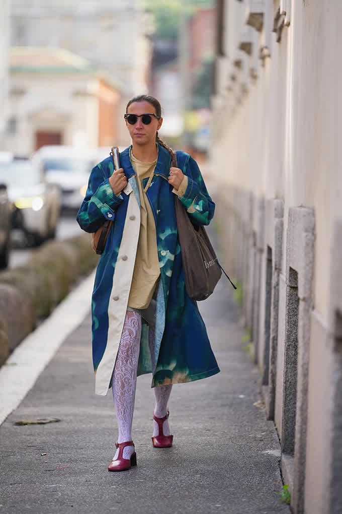 Person in einem blau-grünen Tie-Dye-Mantel mit auffälligen Farbfeldern, kombiniert mit Spitzenstrumpfhose und Mary-Janes