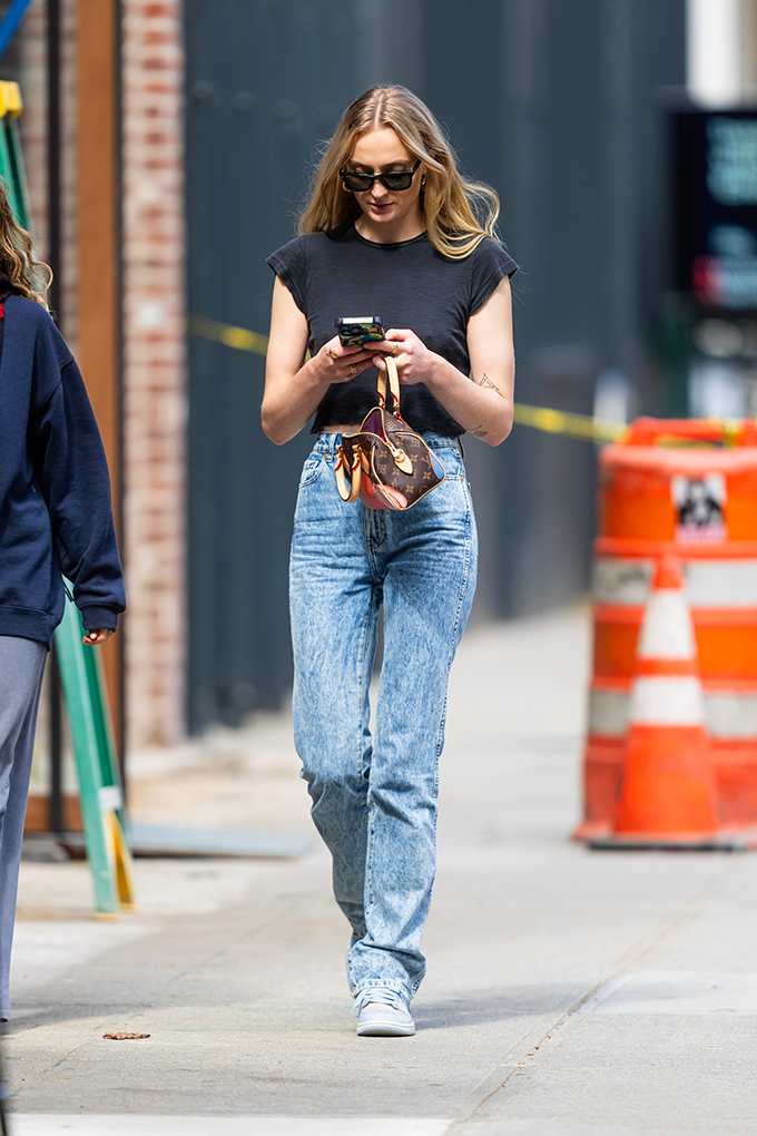Sophie Turner spaziert auf Gehweg, trägt schwarzes Shirt und Jeans, während sie auf Handy tippt