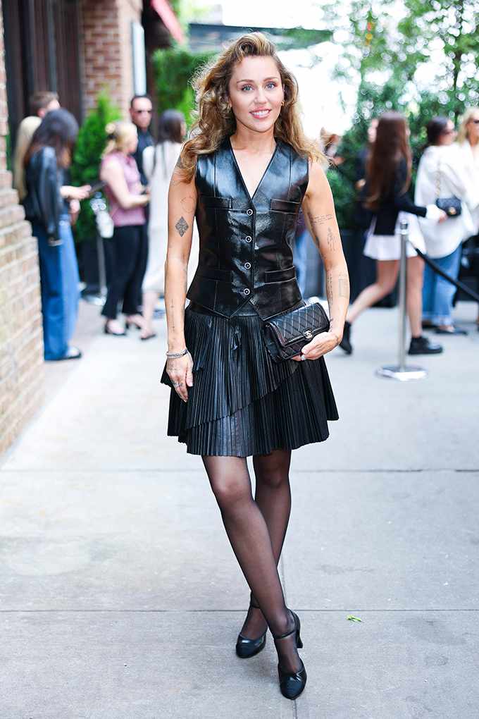 Miley Cyrus in schwarzer Lederweste und Plisseerock trägt Mary Janes mit Blockabsatz und Steppclutch