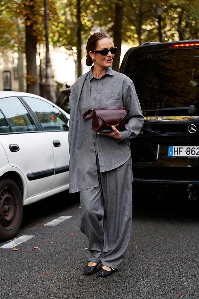 Frau im grauen Layering-Look mit Oversize-Hemd und Marlenehose trägt schwarze Ballerinas und Clutch
