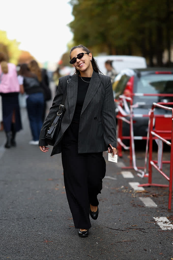 Frau im grauen Nadelstreifen-Blazer und schwarzer Marlenehose trägt Ballerinas mit Schleife im Soft Tailoring-Style