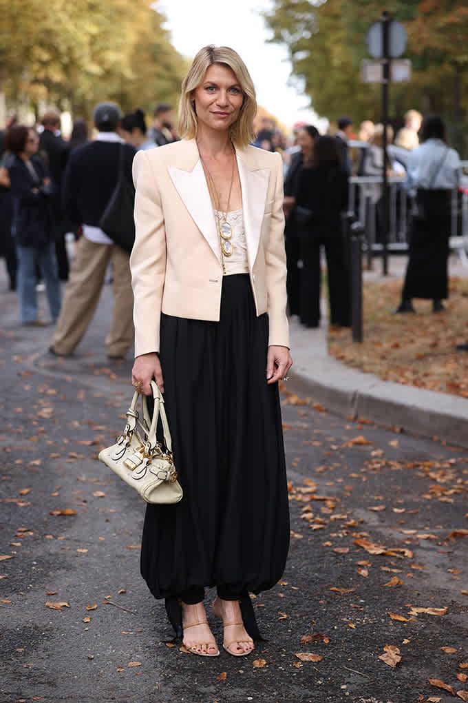 Frau in cremefarbenem Blazer und schwarzer Ballonhose trägt goldene Riemchen-Sandalen und helle Handtasche