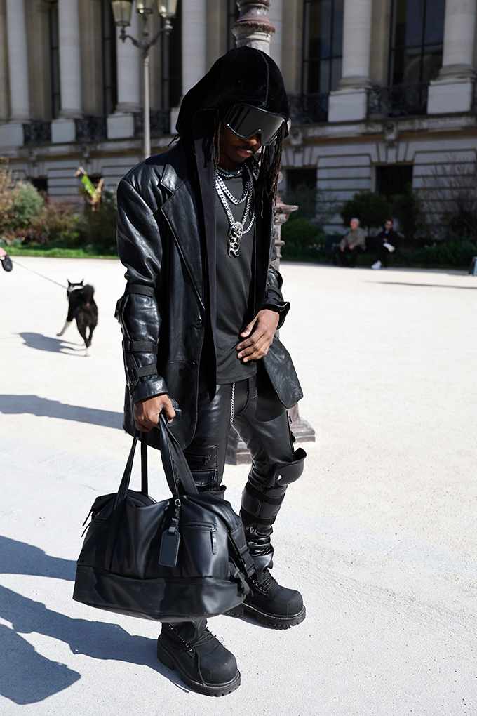 Mann im All-Black-Leder-Look mit Oversize-Jacke und Visierbrille trägt Plateau-Boots und Weekender