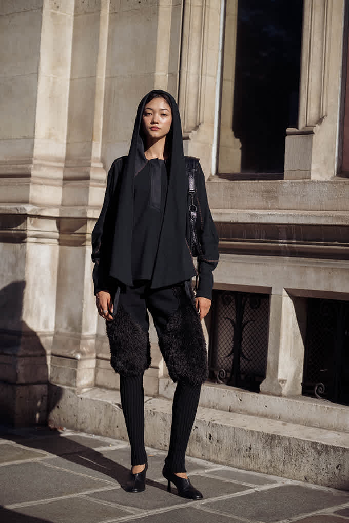 Frau im schwarzen Techwear-Look mit Kapuze, Fellshorts und Beinstulpen trägt Mules mit Stilettoabsatz