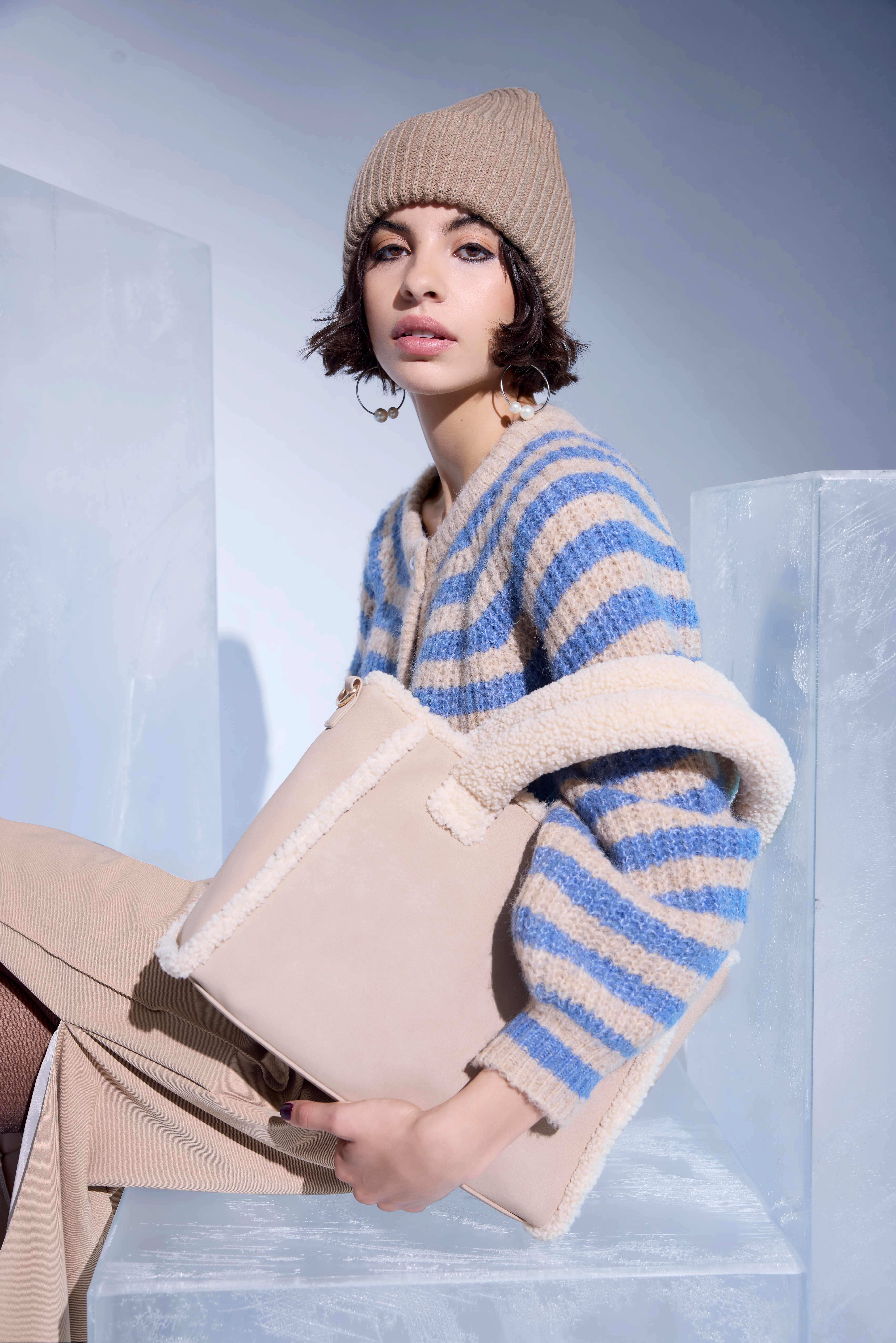 Sitzende Frau mit gestreiftem, flauschigen Pullover und Mütze, mit großer Handtasche unterm Arm