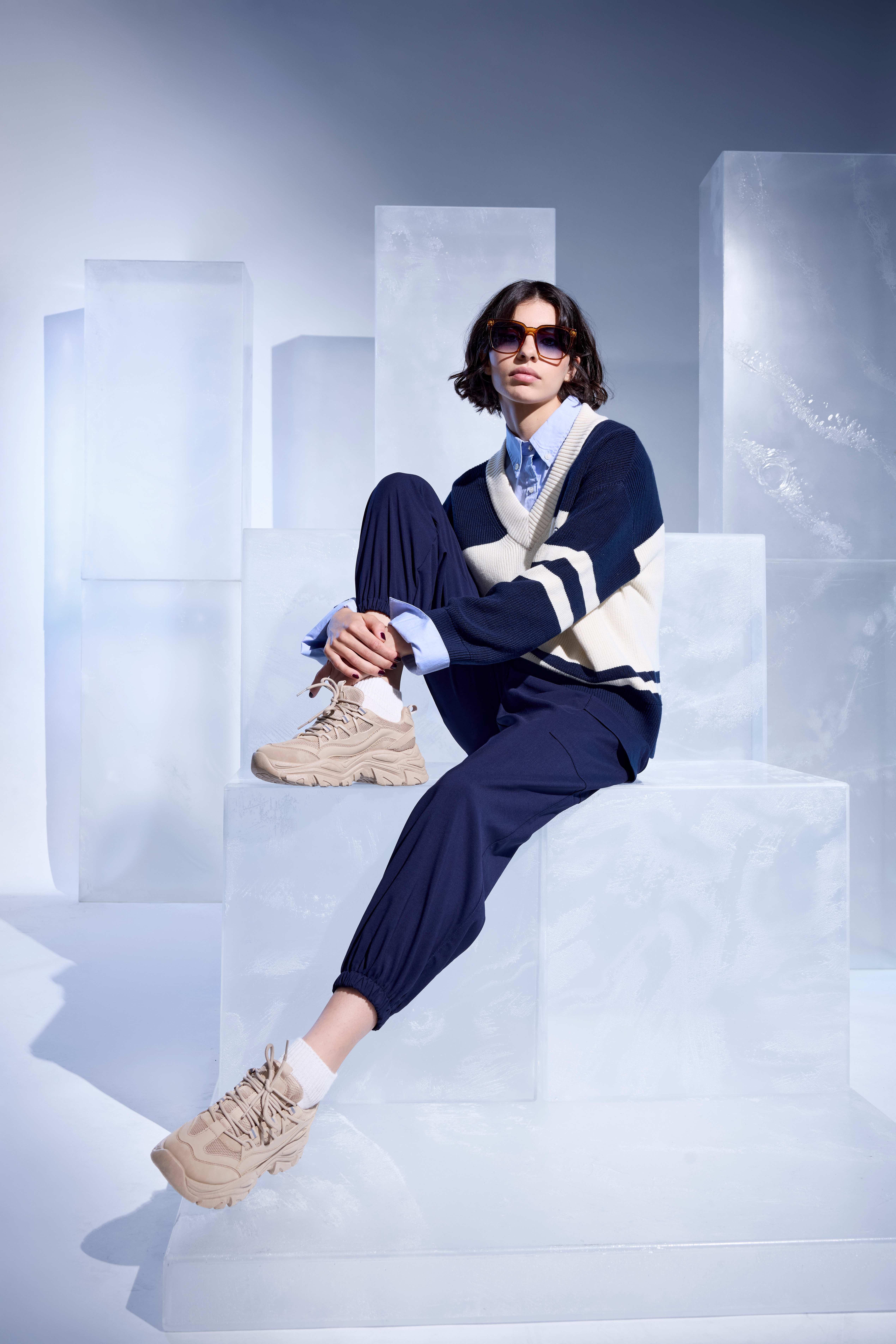 Frau mit beigefarbenen Sneakern, blauer Jogginghose und gestreiftem Pullover, sitzend auf einem transparenten Podest