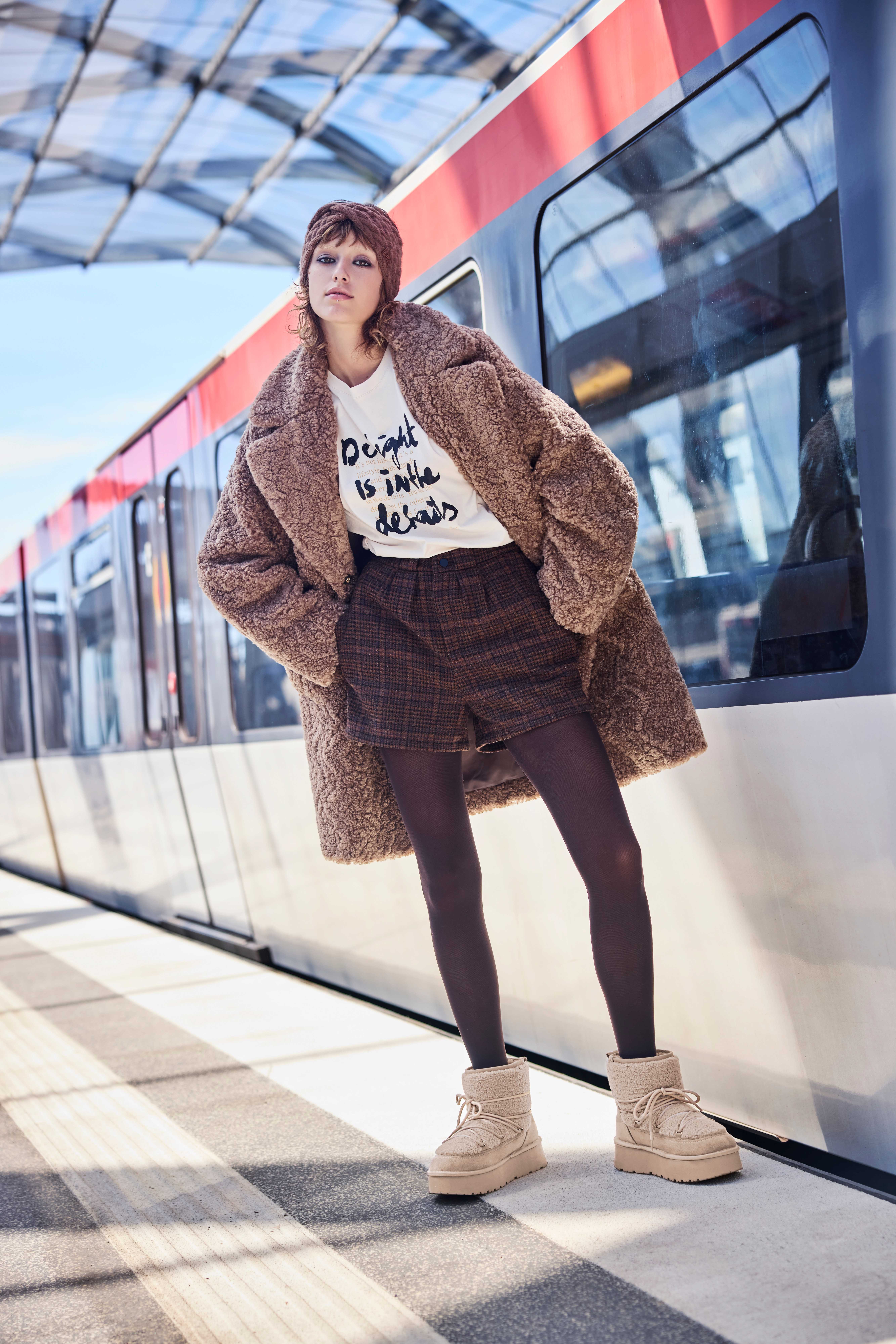 Frau mit Fake Fur-Outfit inklusive Mantel und Mütze, stehend an Bahnsteig vor einem Zug