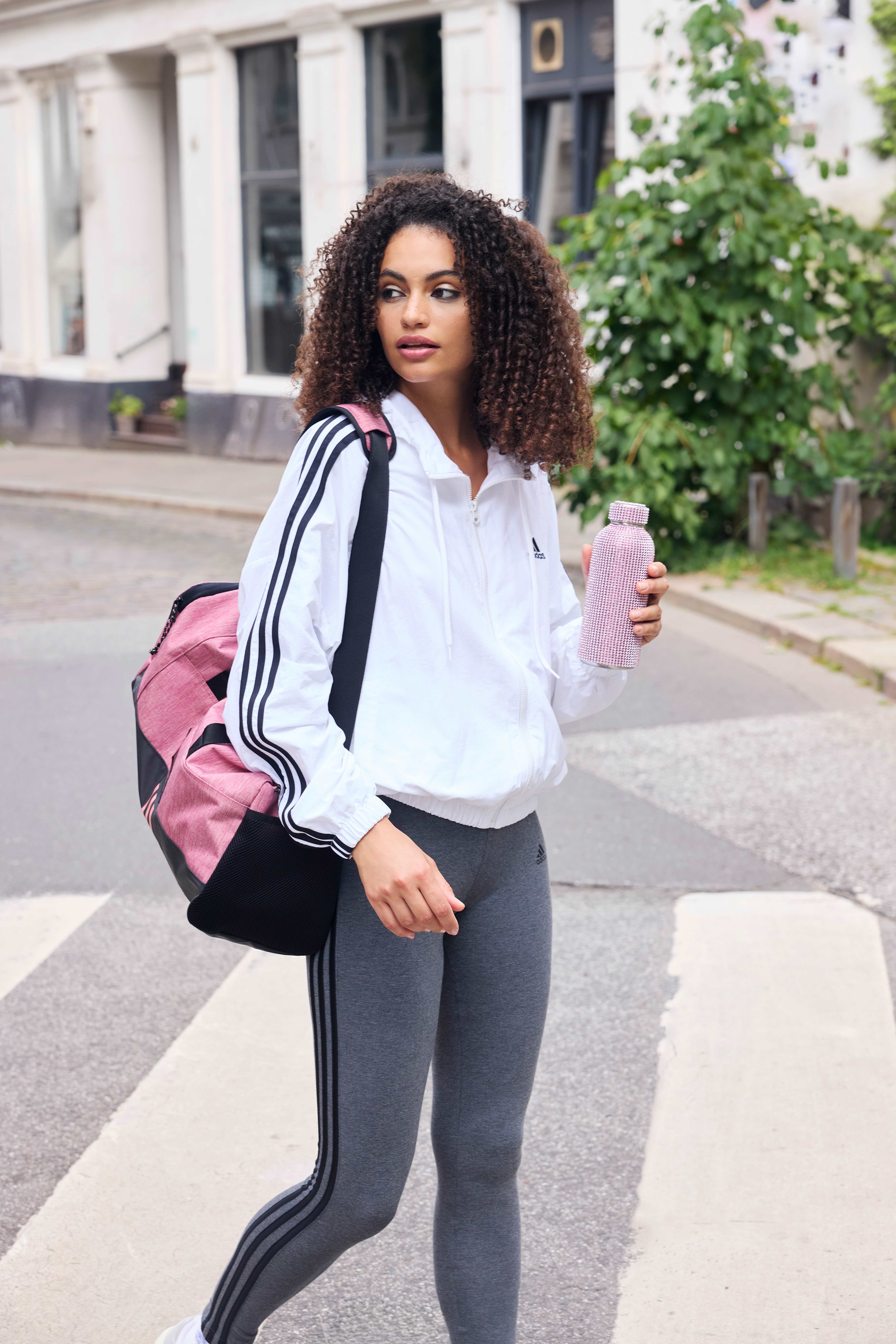 Frau im Athleisure-Look, bestehend aus grauer Leggings und weißer Sportjacke, mit Sporttasche über Schulter