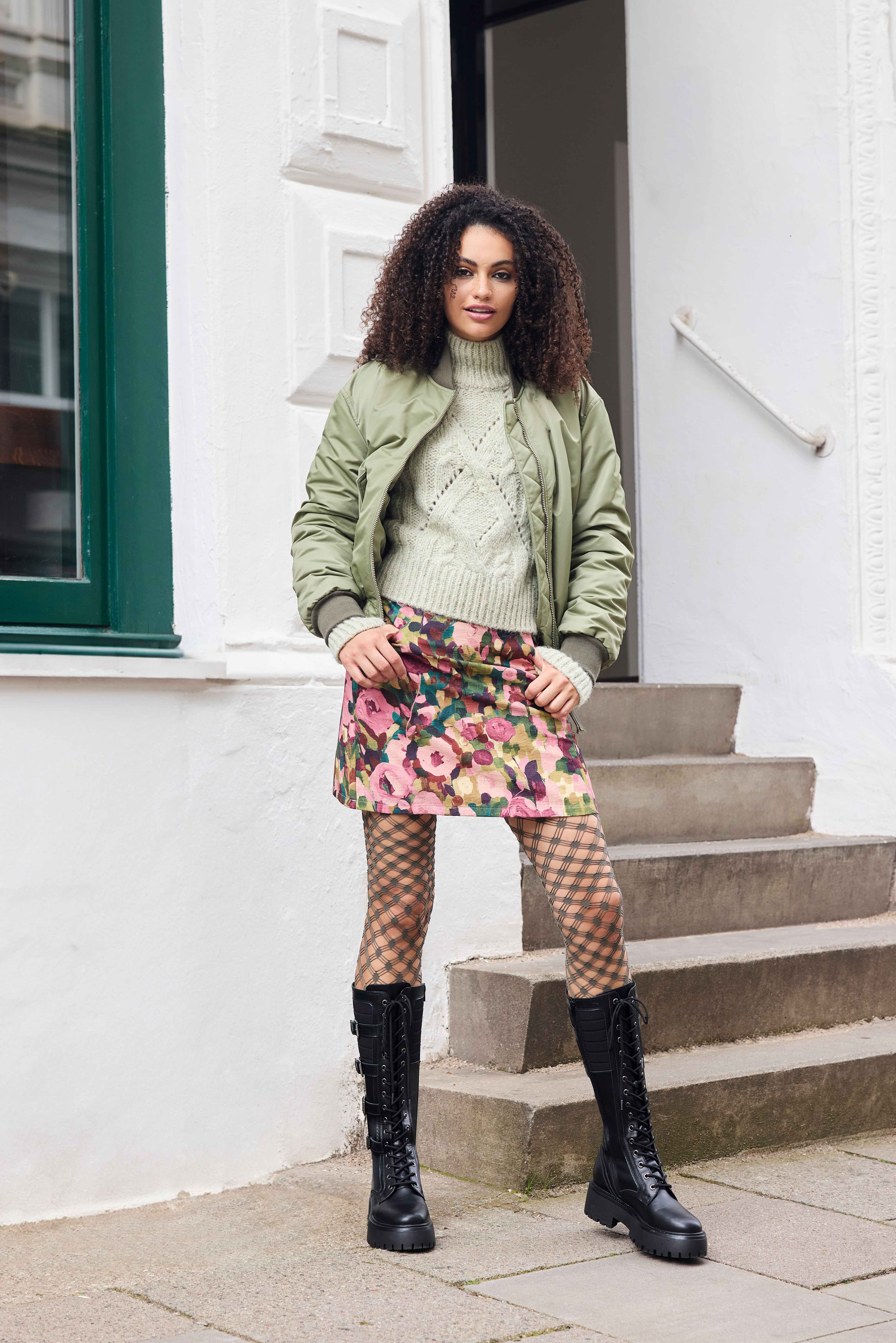 Frau mit grüner Jacke und Top, in Kombination mit Blumenrock und schwarzen Underknees