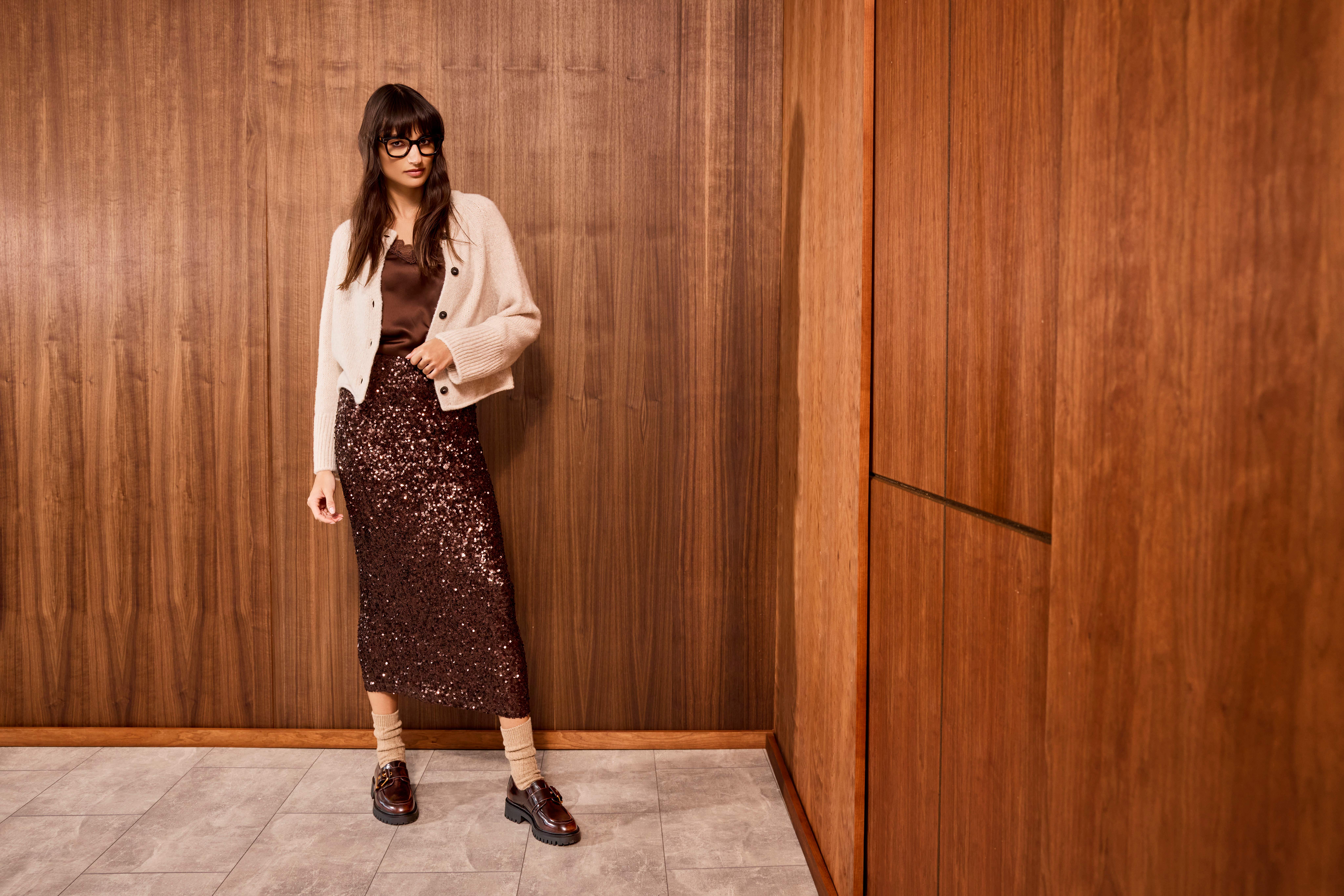 Una donna indossa un top di raso marrone, una gonna marrone ricoperta di paillettes scintillanti e un cardigan beige con mocassini marroni