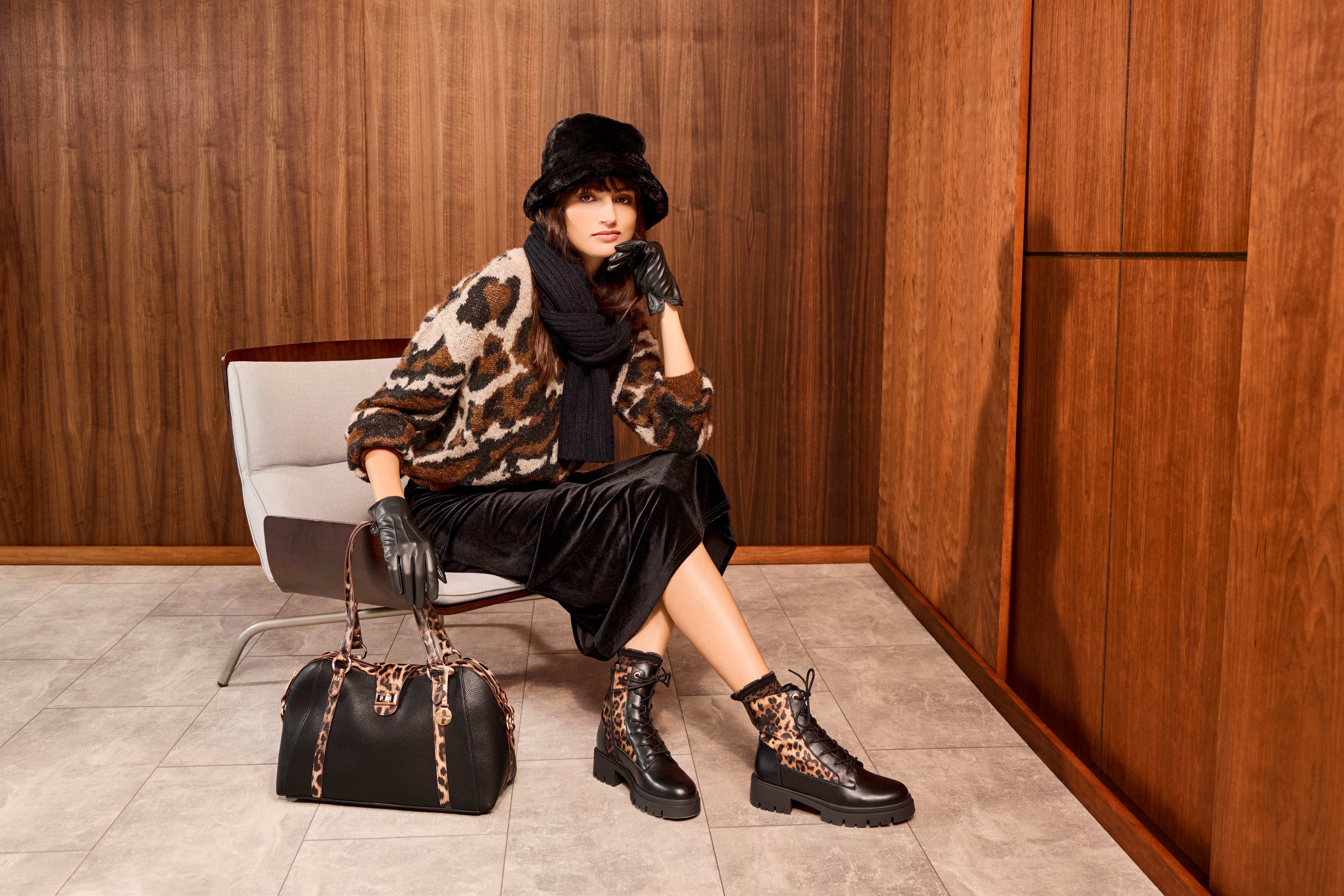 Donna seduta con cappello nero, maglia leopardata e gonna nera, indossa guanti e stivaletti mentre accanto a lei c’è una borsa nera, su sfondo di parete in legno.