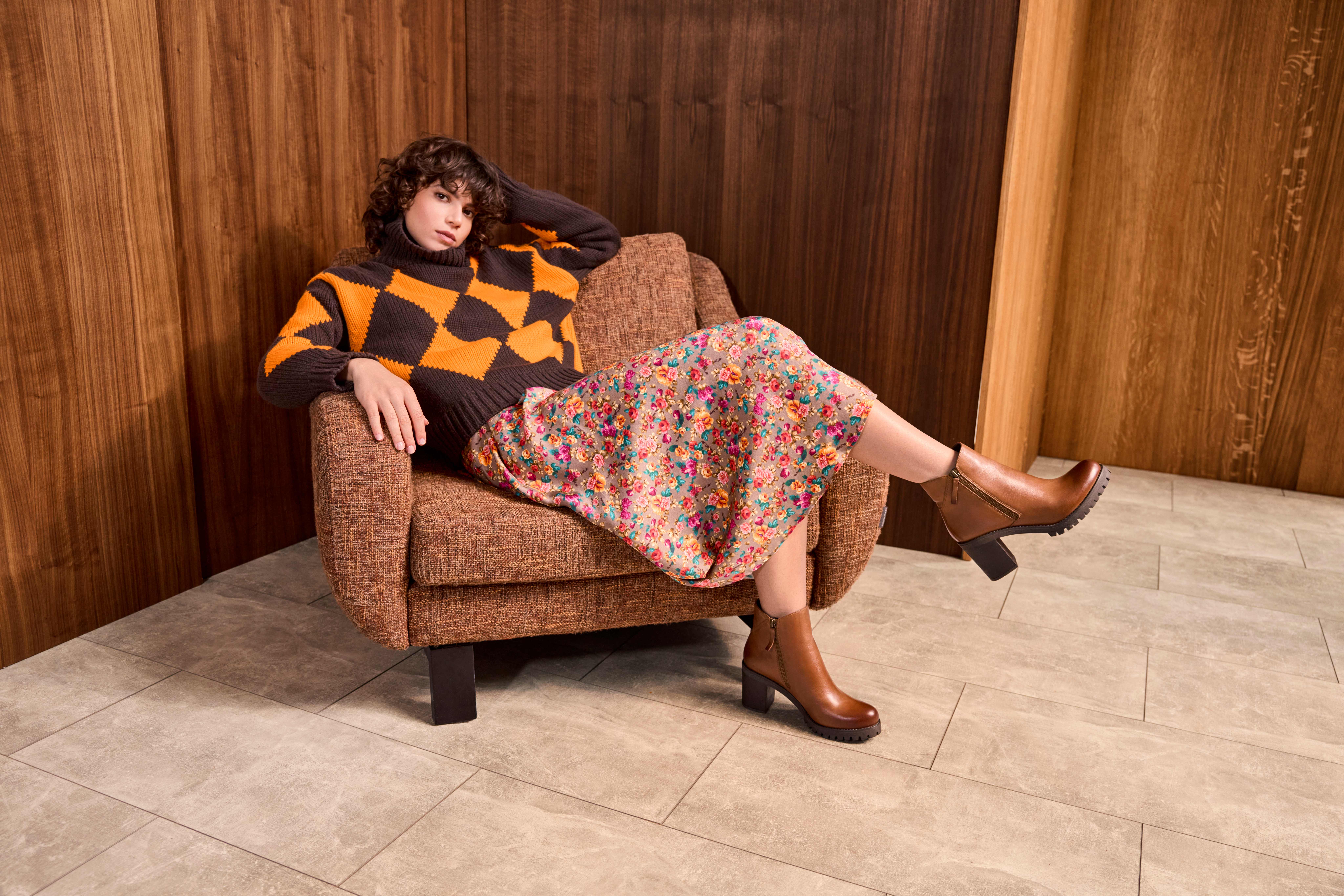 Frau sitzt entspannt in kariertem orange-braunen Pullover mit braunen Ankle Boots auf Sessel