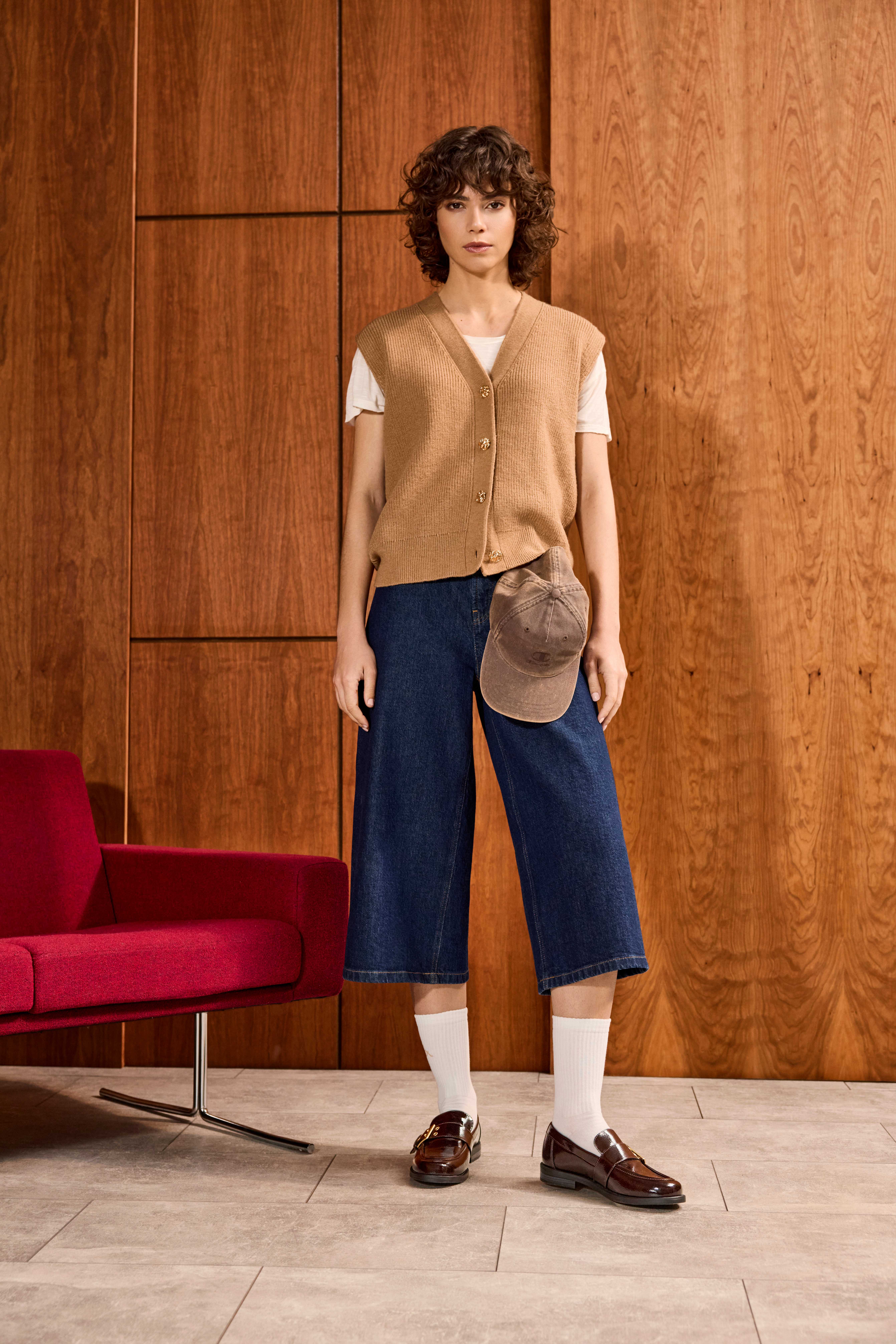 Frau steht lässig, trägt sandfarbenen Pullunder über Shirt, kombiniert mit Denim-Culottes