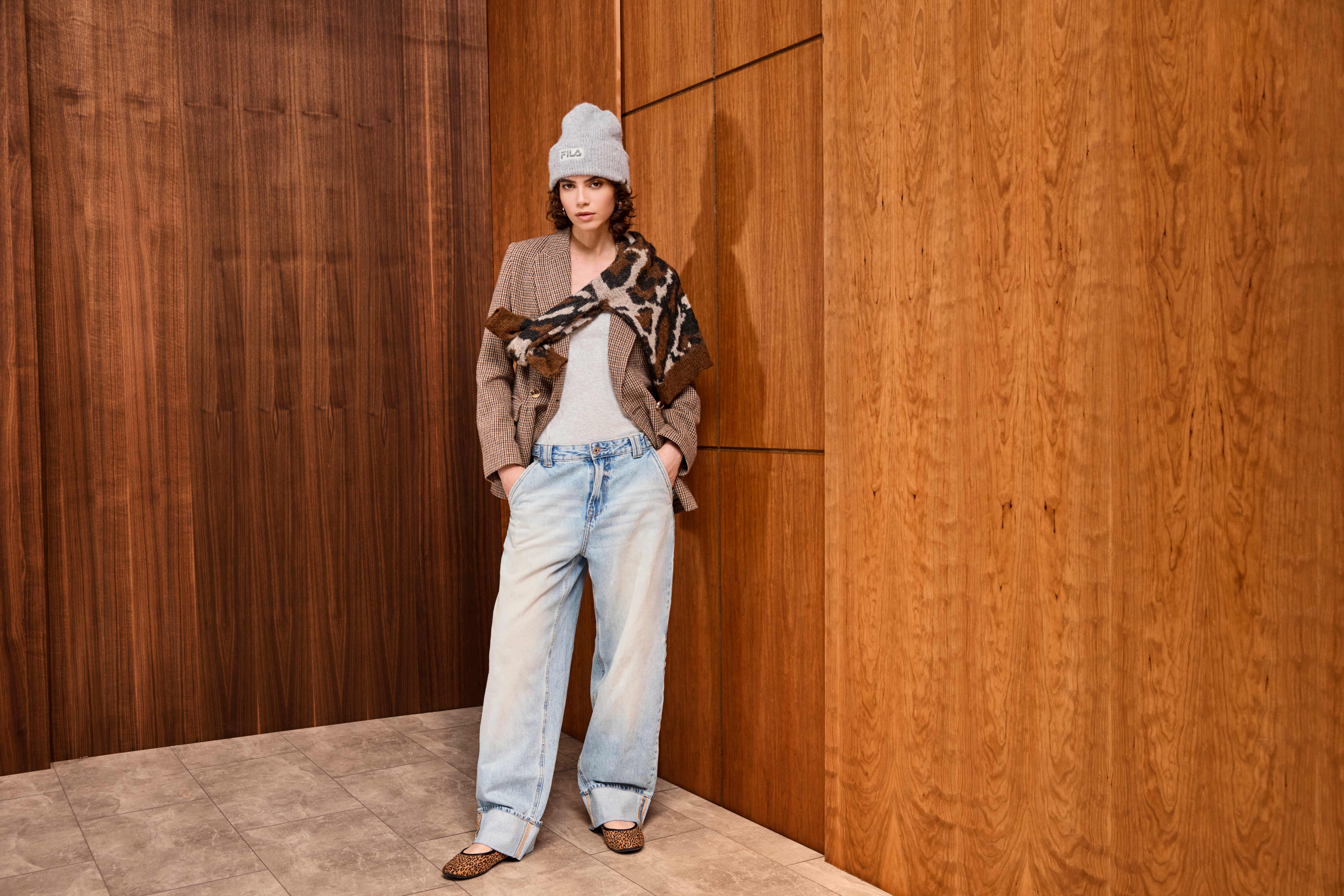 Donna con berretto grigio e capelli ricci appoggiata a una parete in legno, indossa una giacca marrone, jeans larghi, top chiaro e una sciarpa animalier.