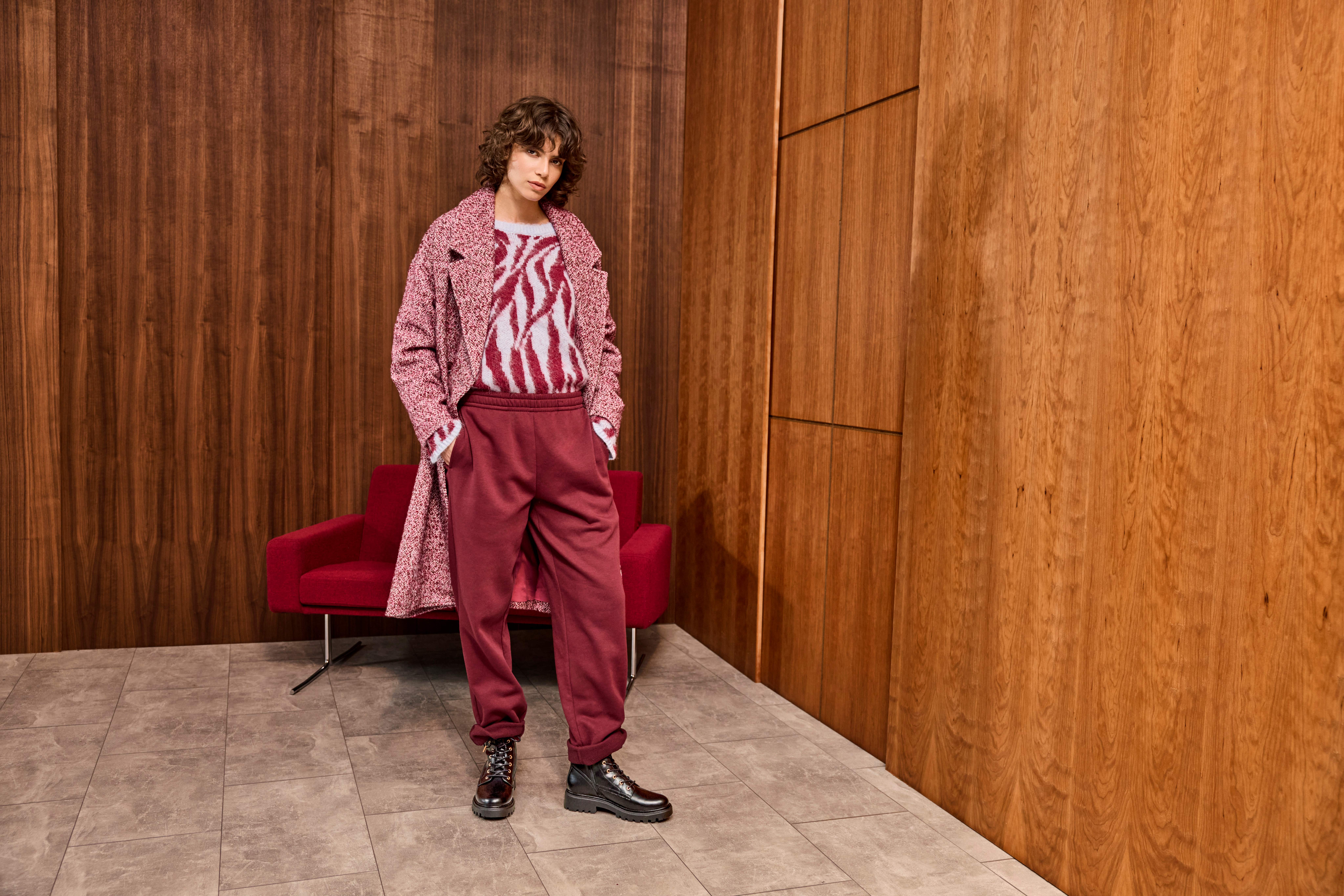 Donna in piedi in un interno moderno con pareti in legno, indossa un cappotto rosa testurizzato, maglione con motivo zebra, pantaloni bordeaux e stivali neri; dietro di lei una poltrona rossa.