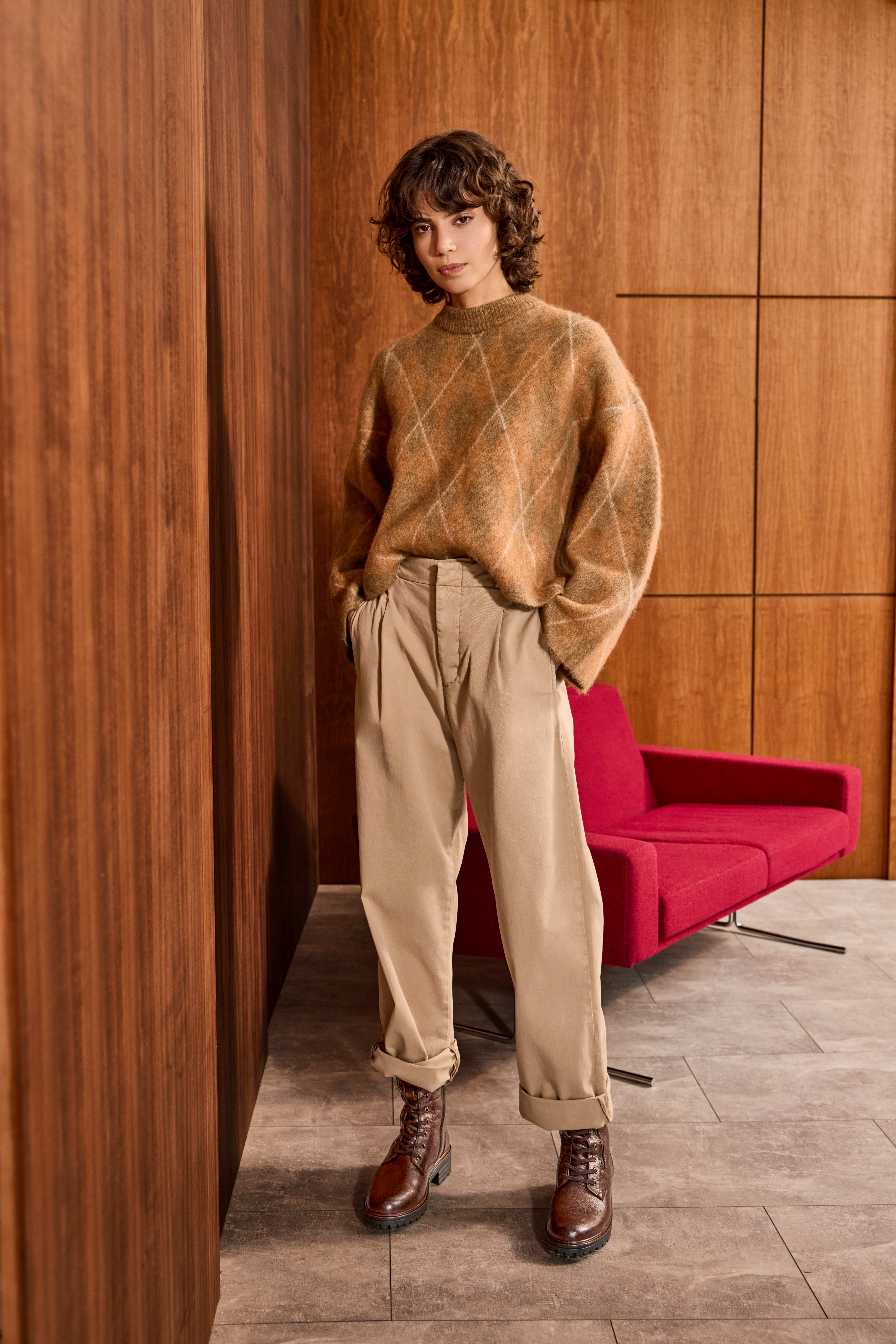Frau mit braunem Pullover, grauer Hose und Lederstiefeln steht vor rotem Sofa