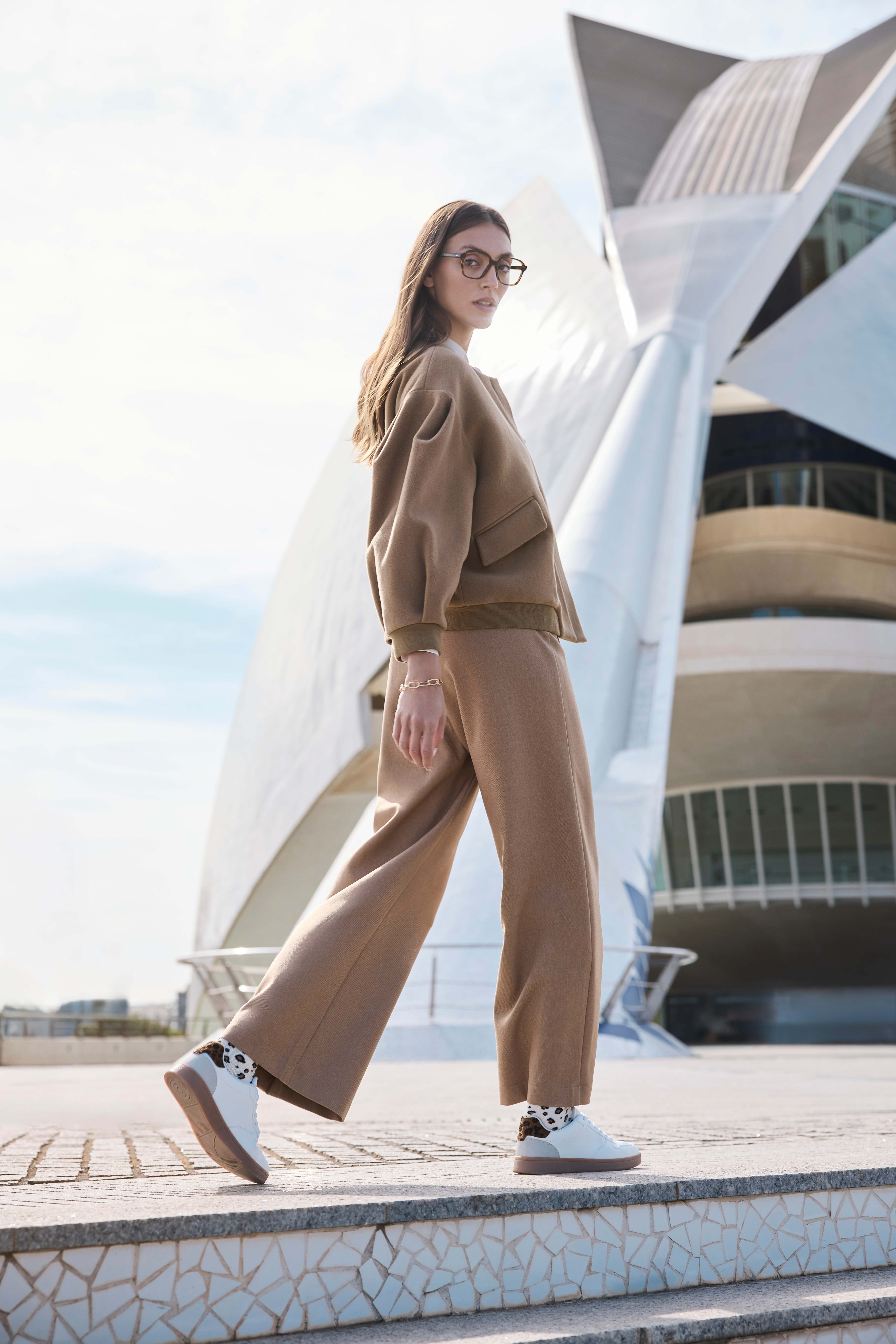 Eine junge Frau trägt eine hellbraune Jogginghose, eine hellbraune Jacke und weiße Sneaker