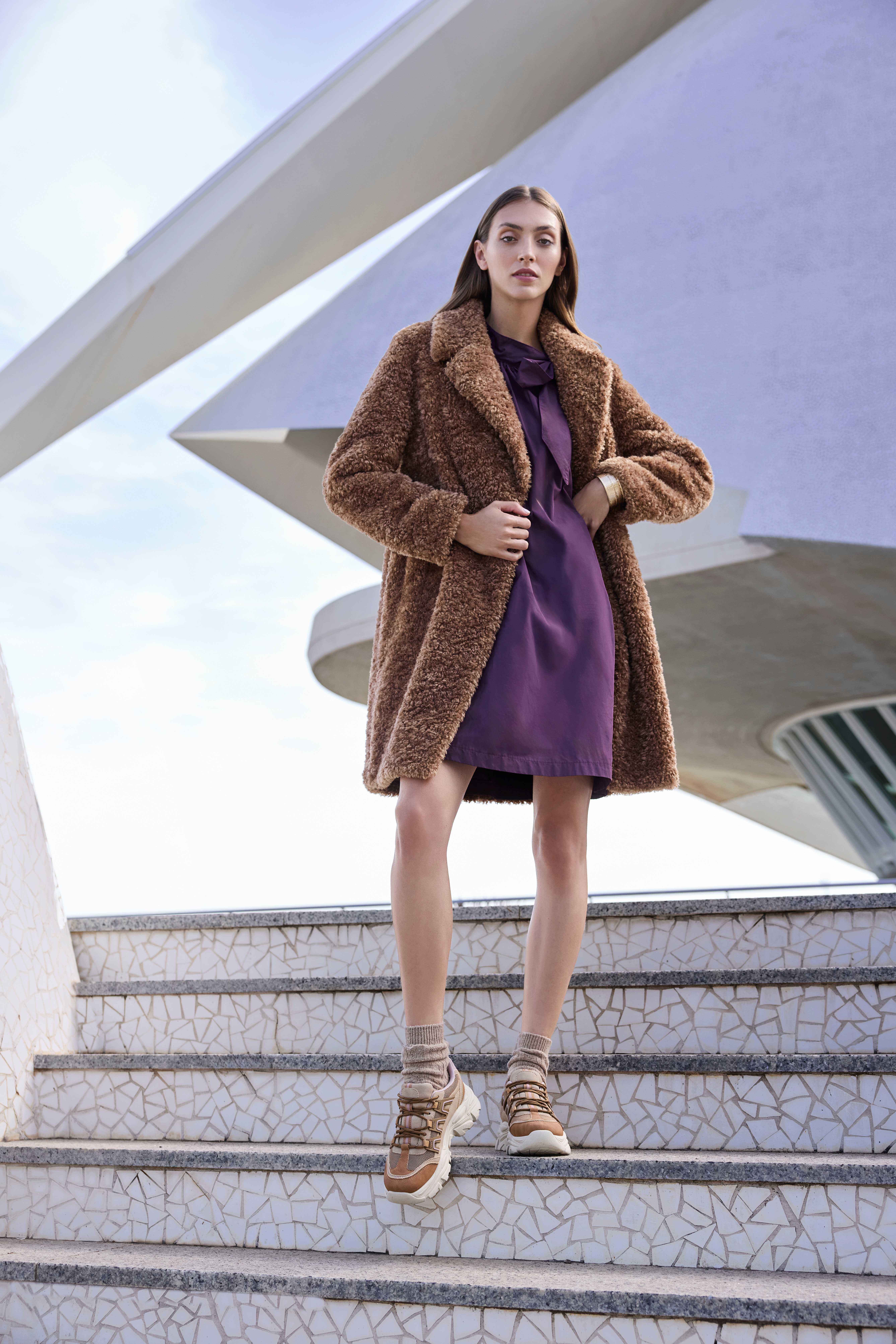 Eine Frau trägt einen braunen Teddy-Mantel, ein lila Kleid und beige Sneakers mit Socken