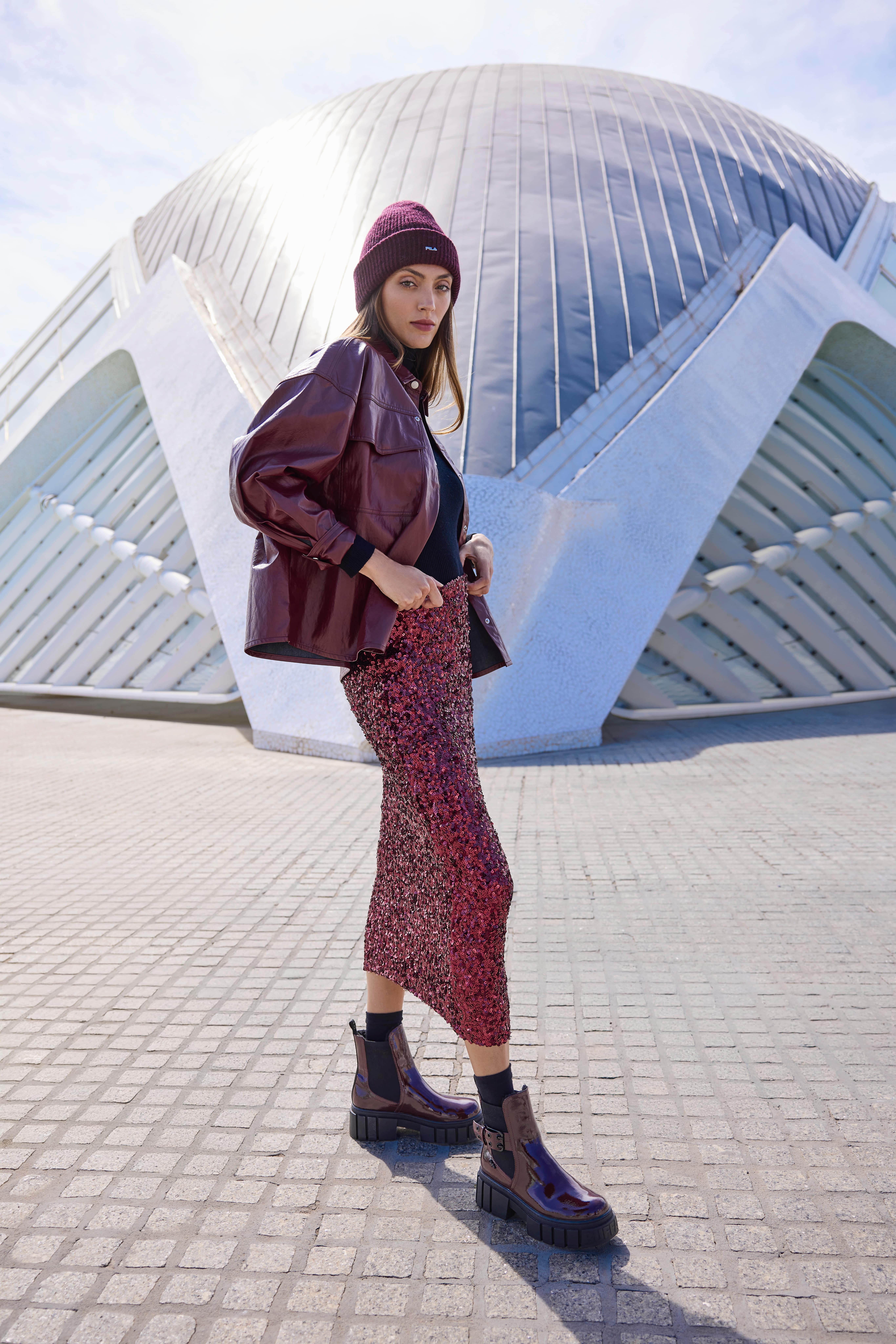 Eine Frau trägt eine burgunderrote Lackjacke, eine passende Mütze und einen glitzernden Rock mit klobigen Stiefeletten