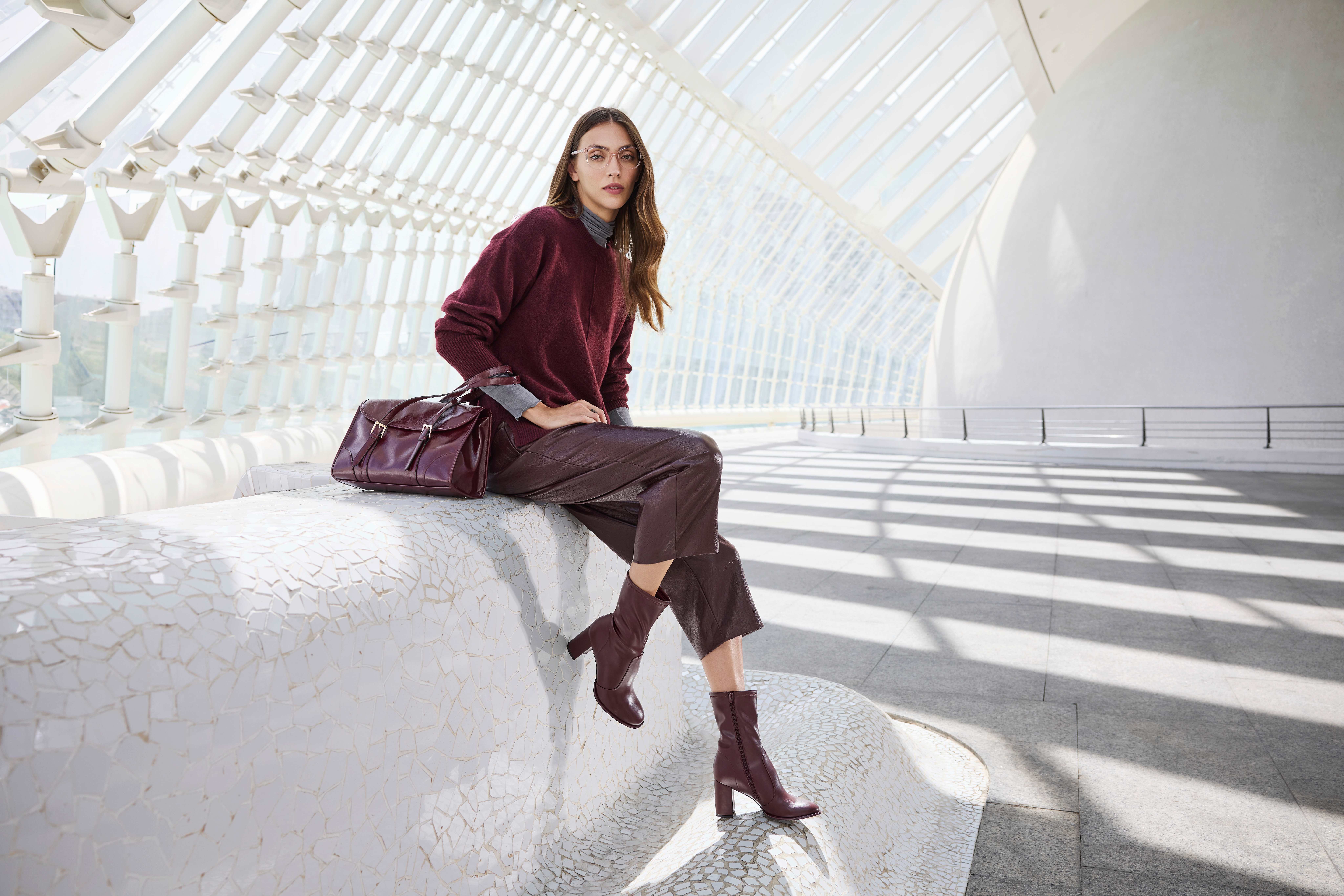 Una donna indossa un maglione bordeaux, pantaloni di pelle abbinati, stivaletti con il tacco e una borsa coordinata