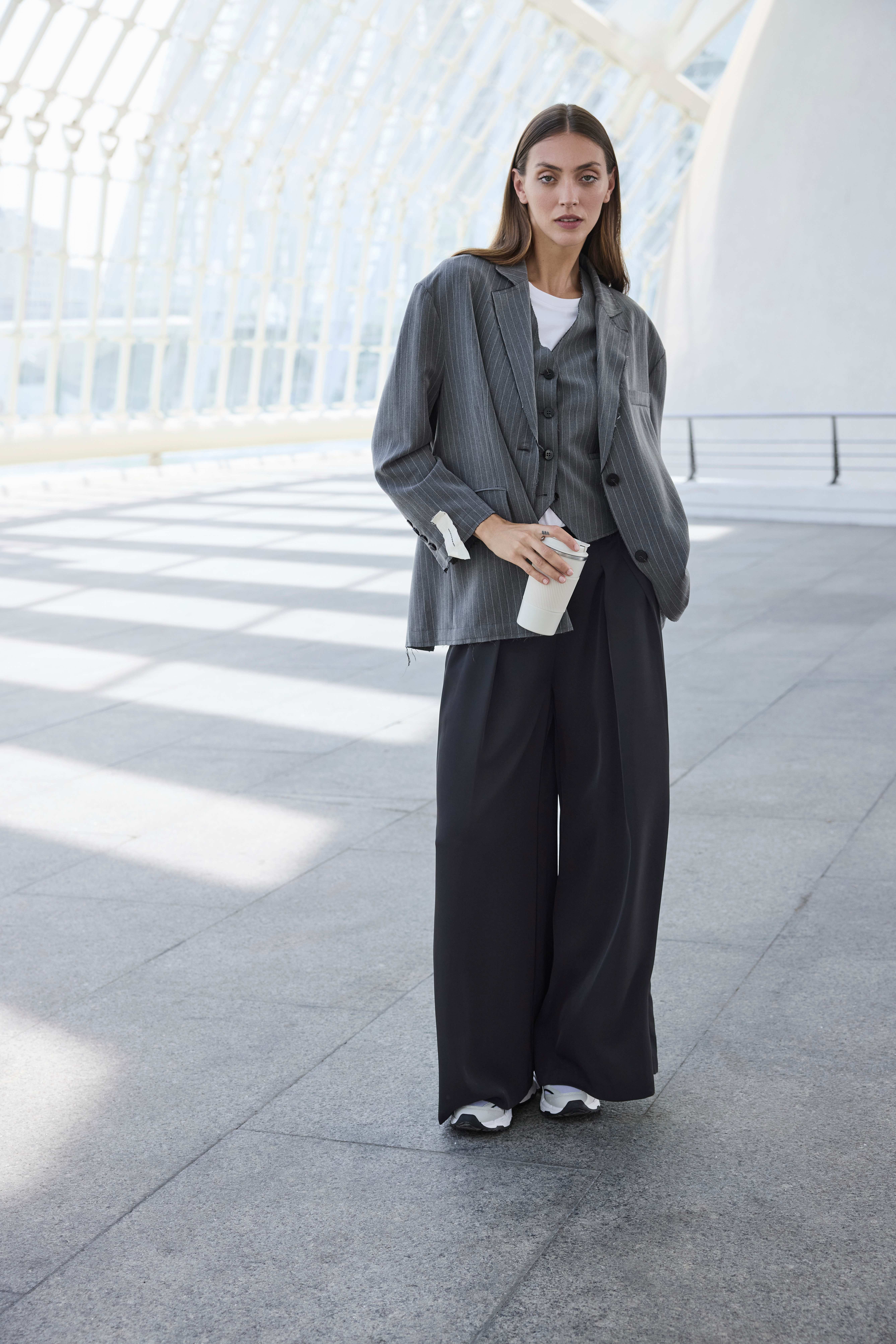 Eine junge Frau trägt eine lockere schwarze Hose, einen grauen Blazer und hält einen hellen Becher in der Hand