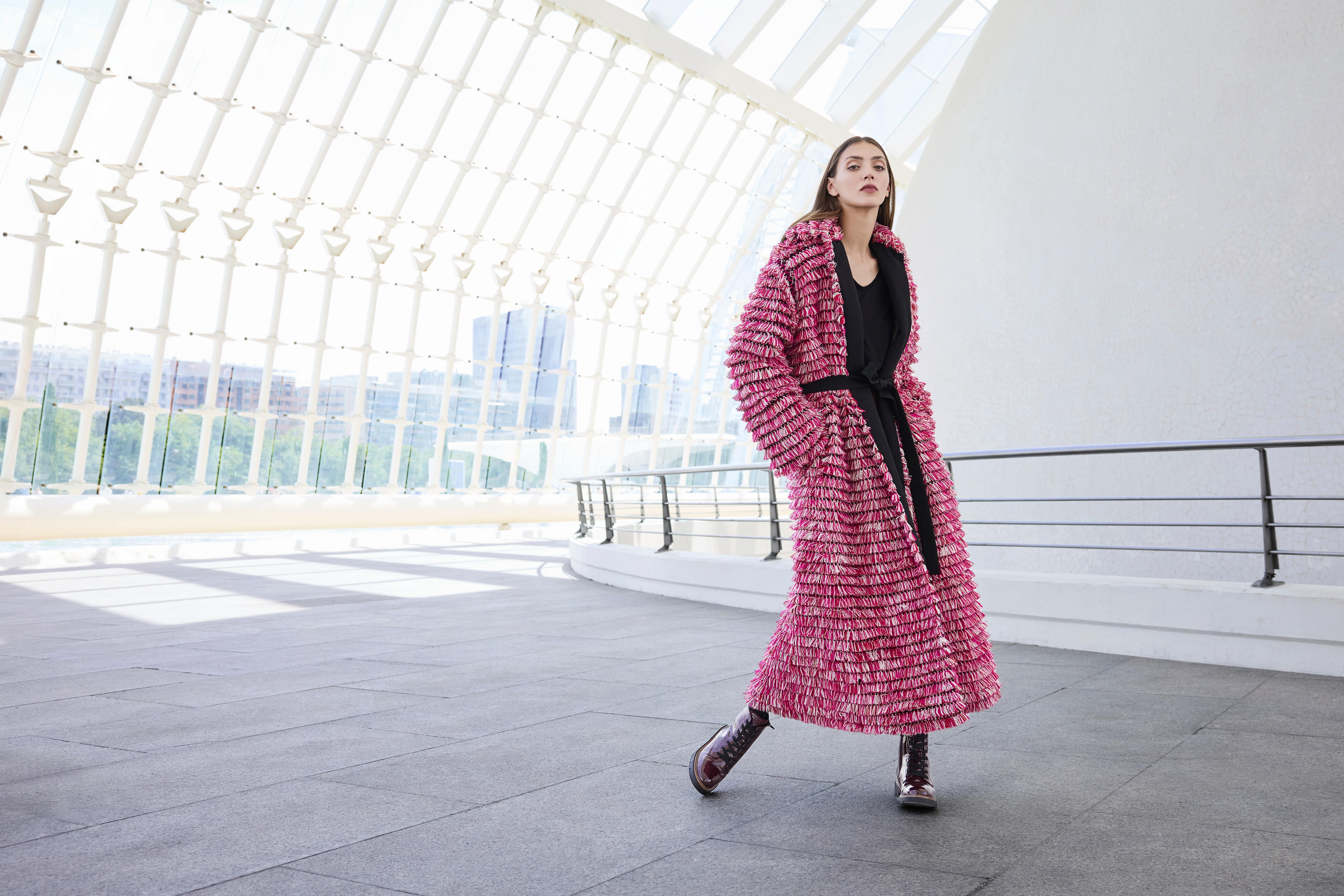 Una donna indossa un cappotto rosa con texture e cintura nera, abbinato a un completo nero e stivaletti stringati rosso scuro