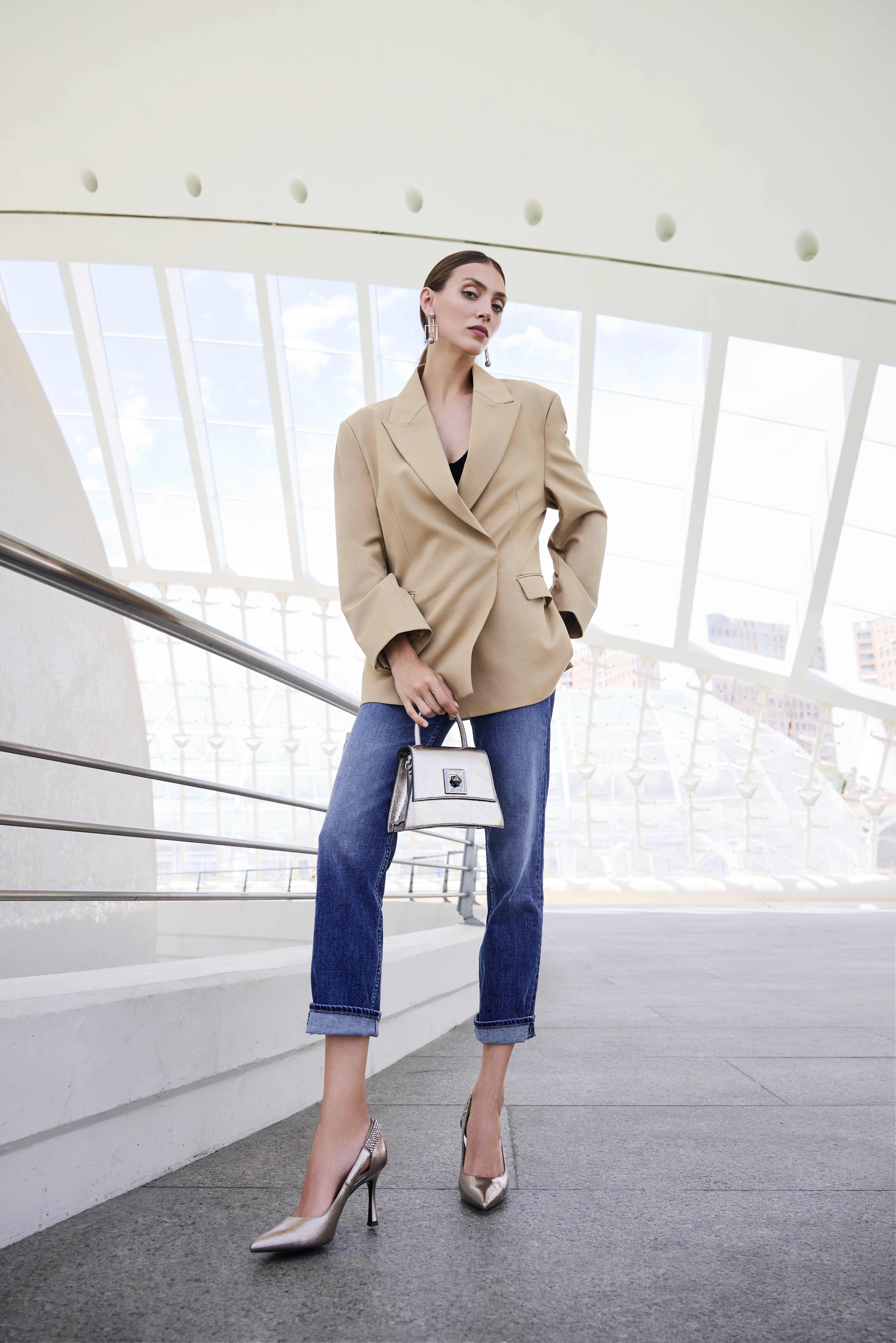 Eine Frau trägt einen beigen Blazer, eine blaue Jeans und silberne hohe Schuhe, in ihrer Hand hält sie eine weiße Tasche