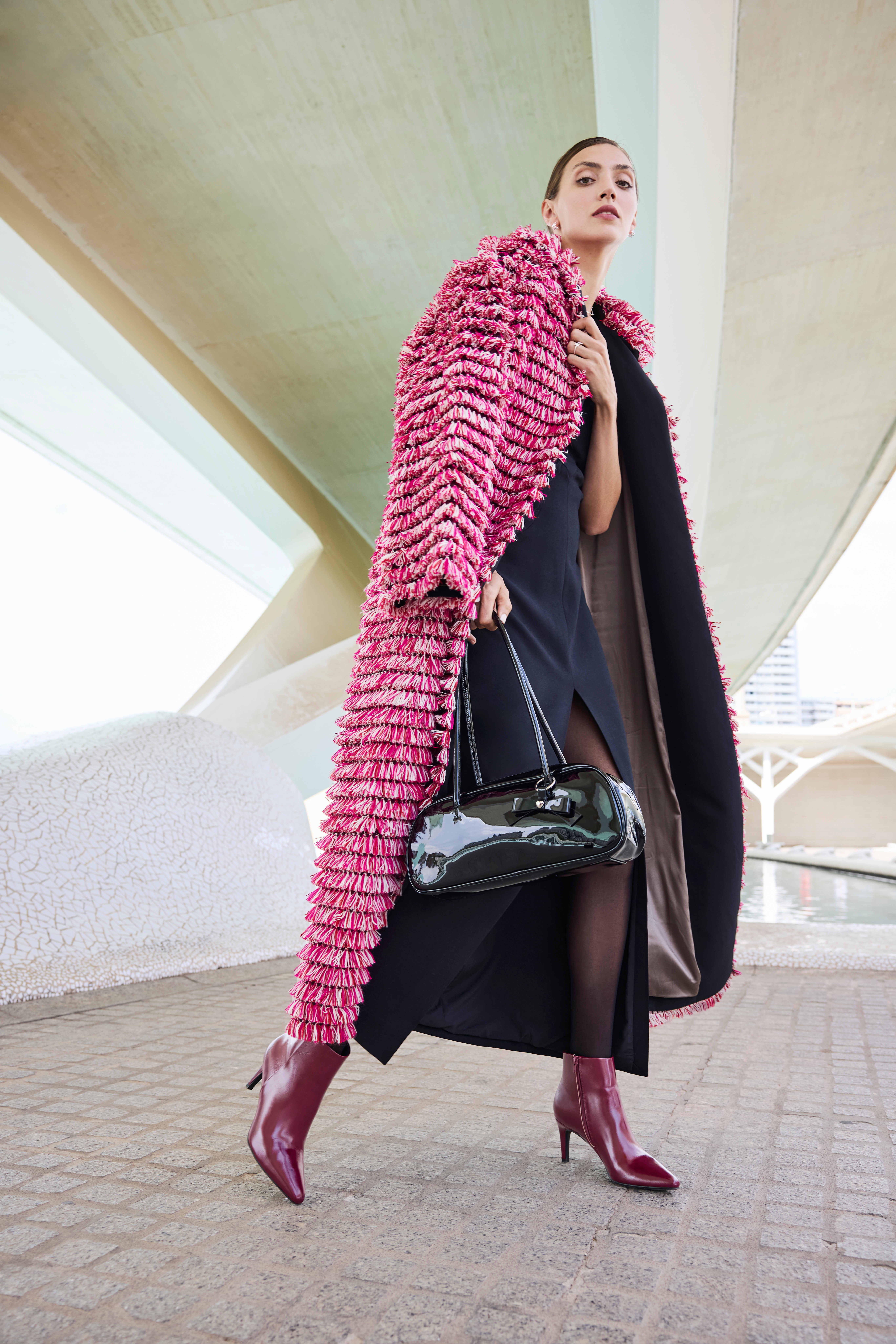 Una donna indossa un elegante abito nero con spacco, abbinato a un lungo cappotto strutturato rosa; completa il look con stivaletti con tacco color bordeaux e una borsa nera lucida