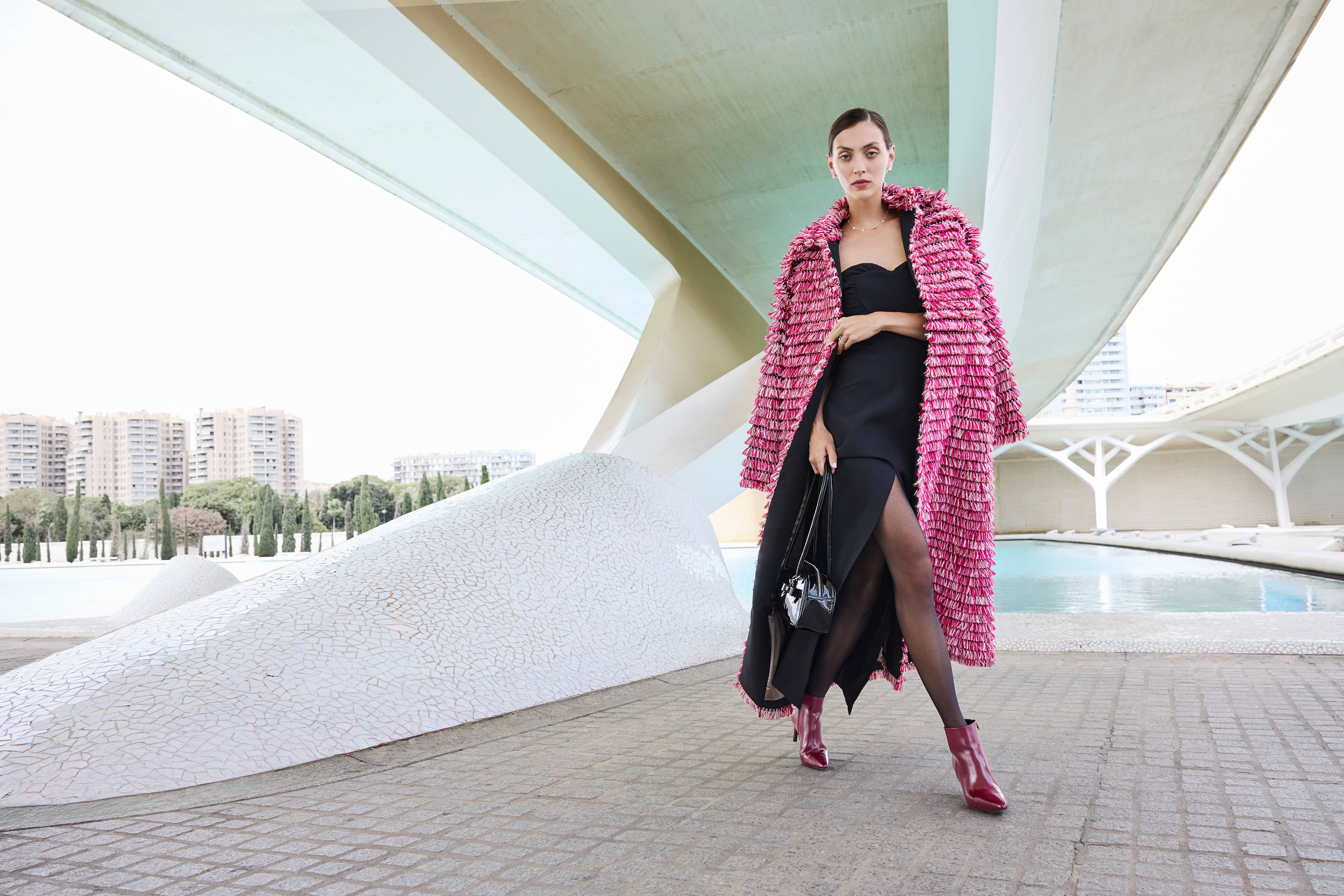 Una donna indossa un abito nero con uno spacco profondo e un vistoso cappotto rosa; abbina il tutto a una borsa nera e a stivaletti bordeaux