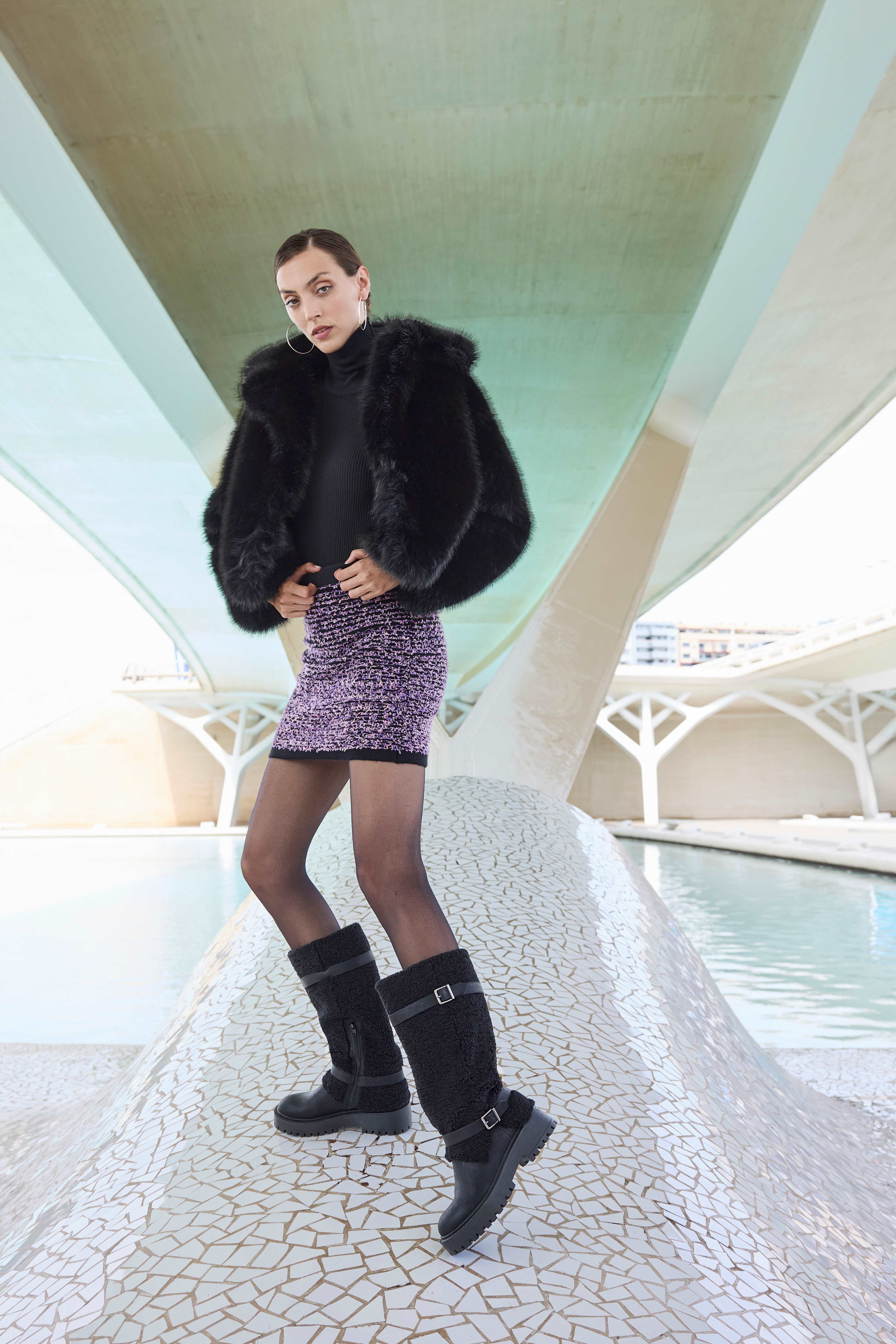Una donna indossa un cappotto di pelliccia sintetica nera, un maglione a collo alto nero, una gonna corta viola, collant neri e stivali neri al ginocchio con fibbie