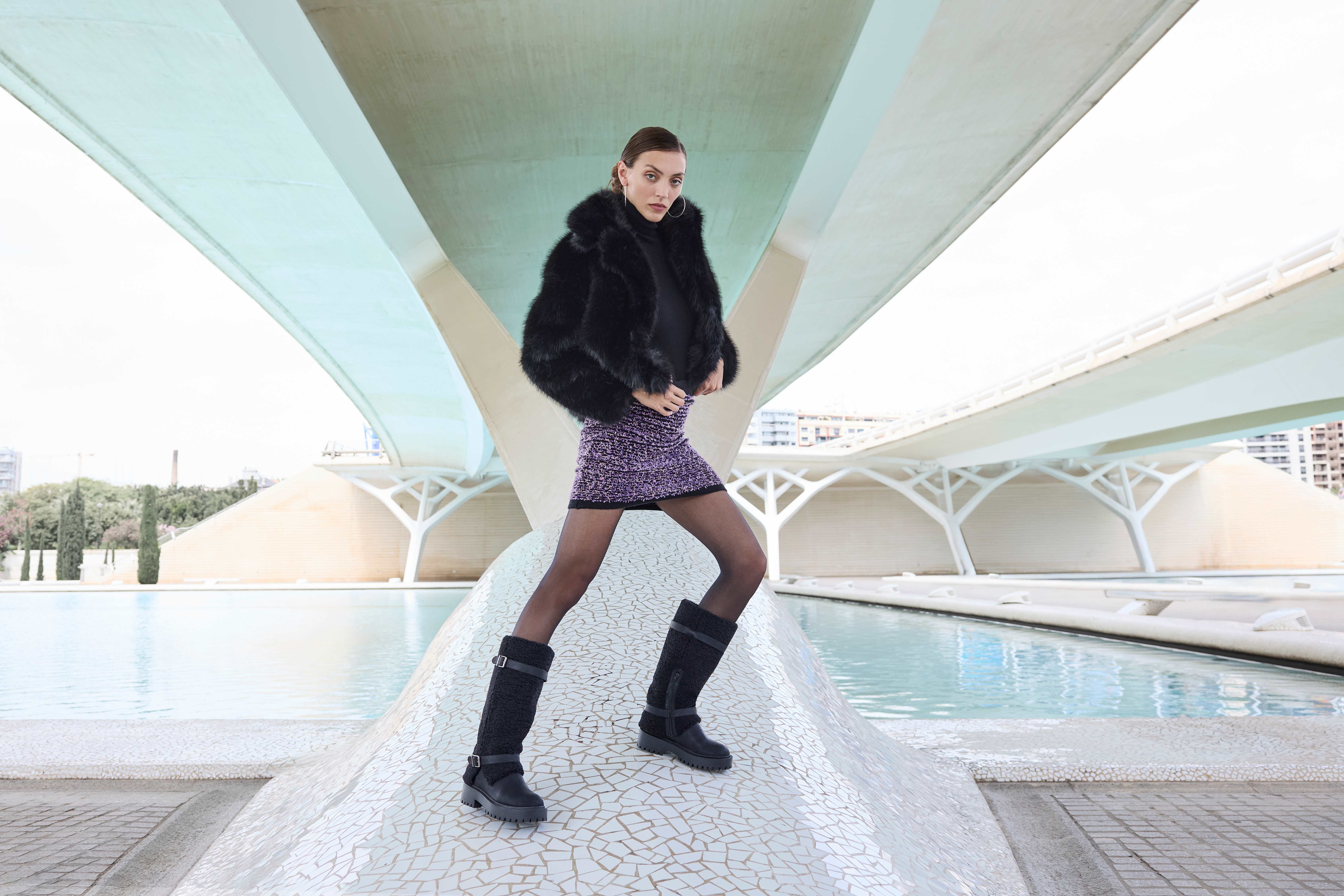 Eine Frau trägt eine schwarze Kunstpelzjacke, einen lilafarbenen Rock, eine schwarze Strumpfhose und schwarze Stiefel