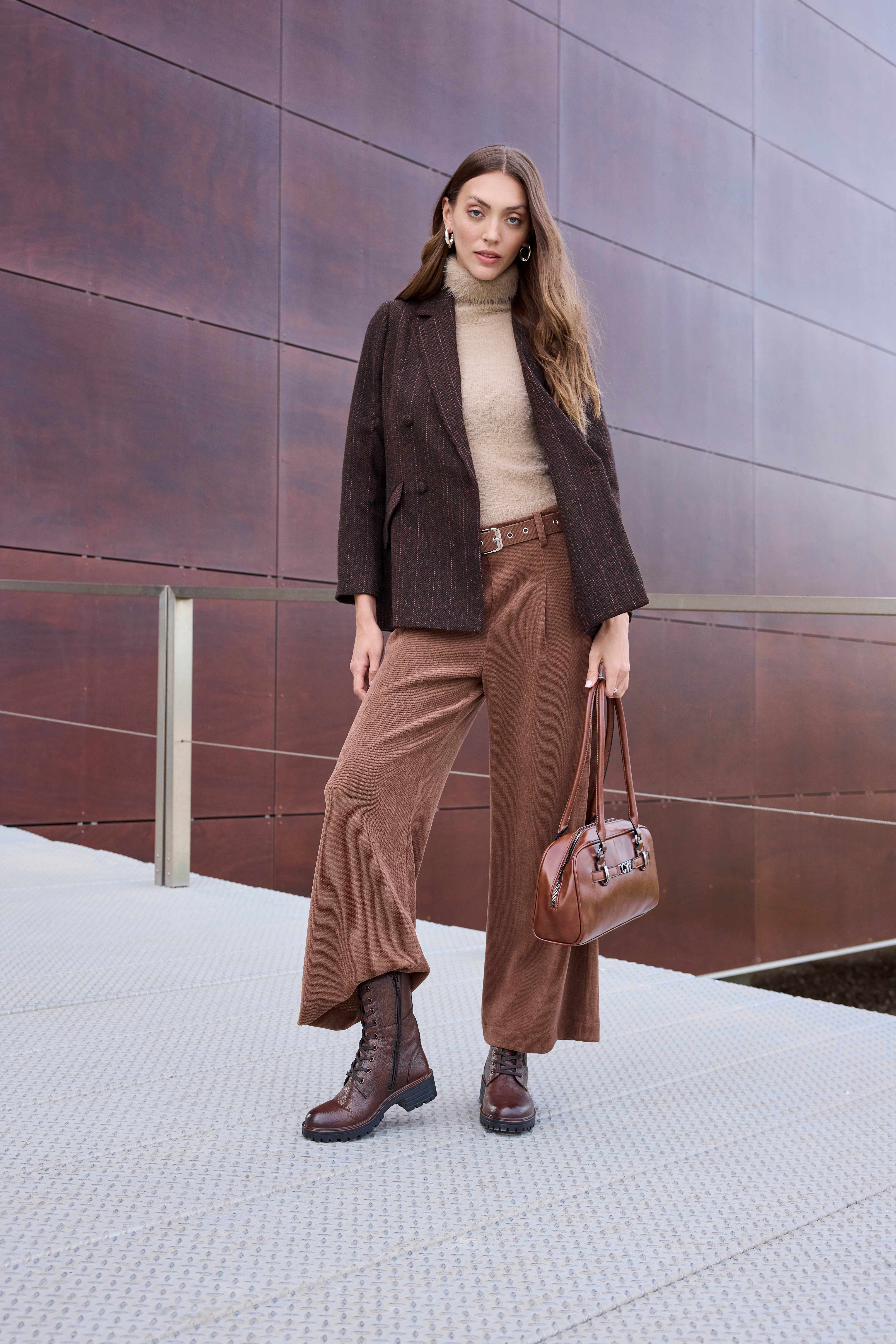 Eine Frau trägt einen braunen Blazer mit Rollkragenpullover, weiter brauner Hose, Schnürboots und einer braunen Lederhandtasche
