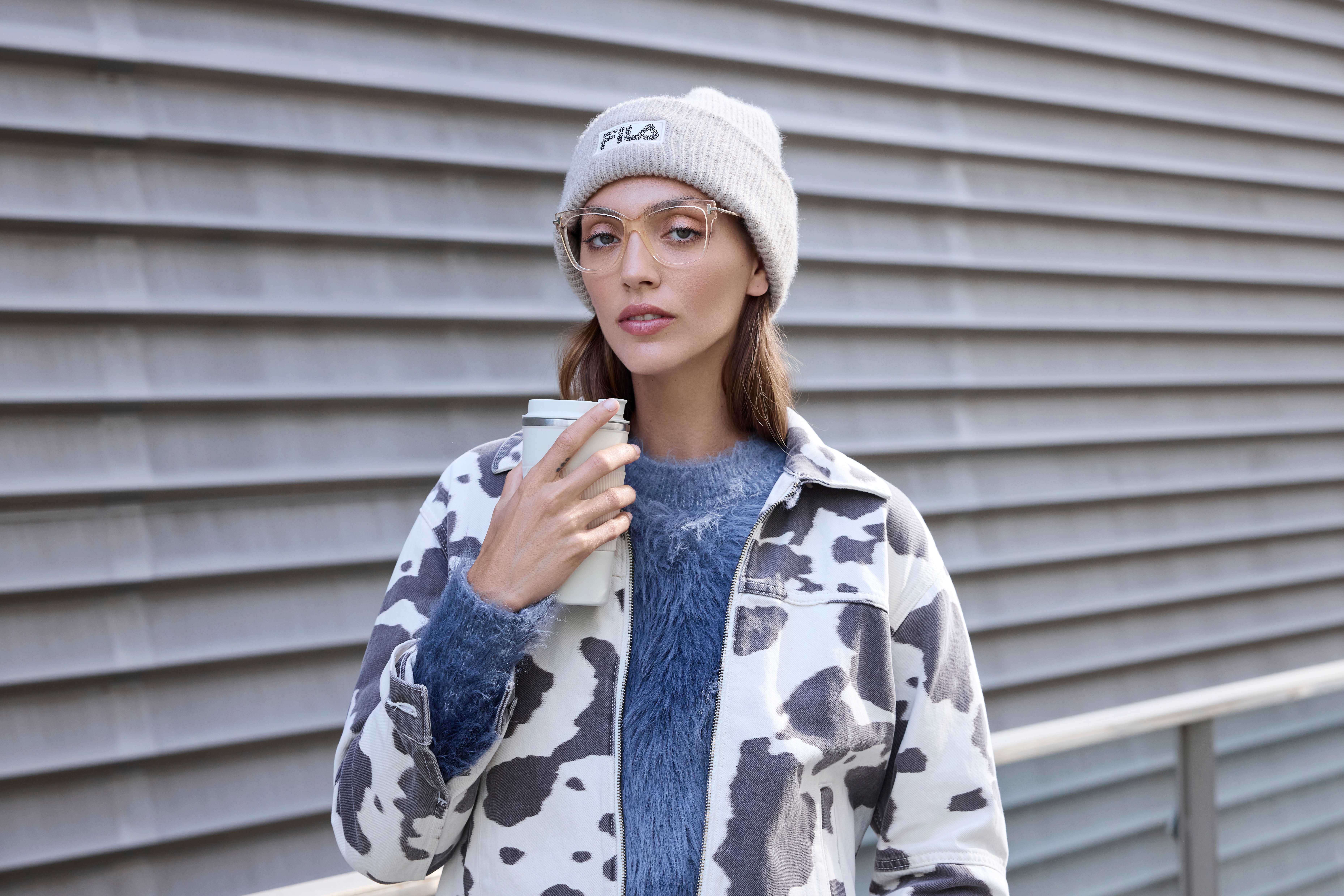 Donna con berretto beige e occhiali tiene una tazza da asporto, indossa un maglione blu e una giacca maculata davanti a una parete a pannelli.