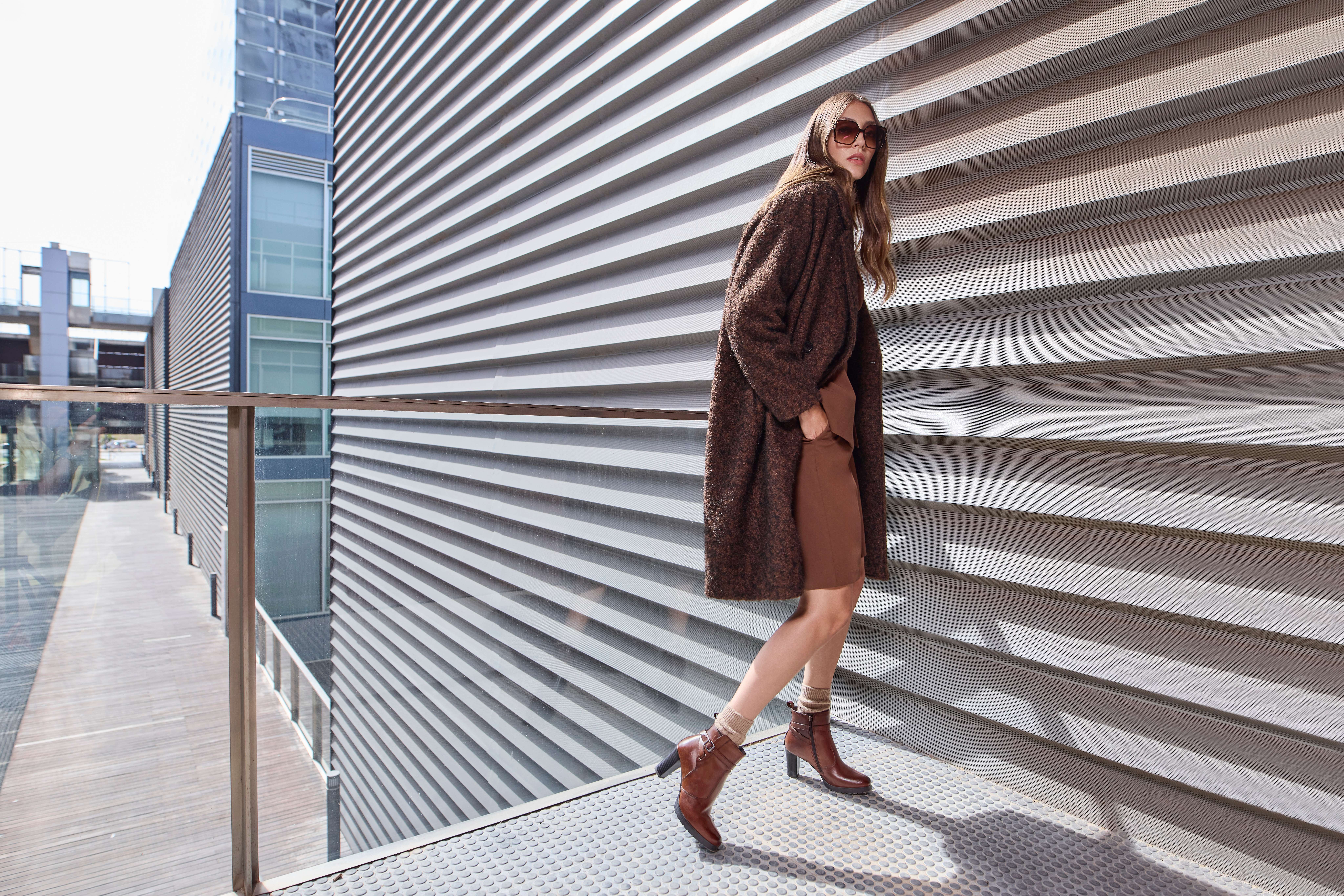 Eine Frau trägt einen braunen Mantel, ein braune kurze Stoffhose und passende Stiefeletten