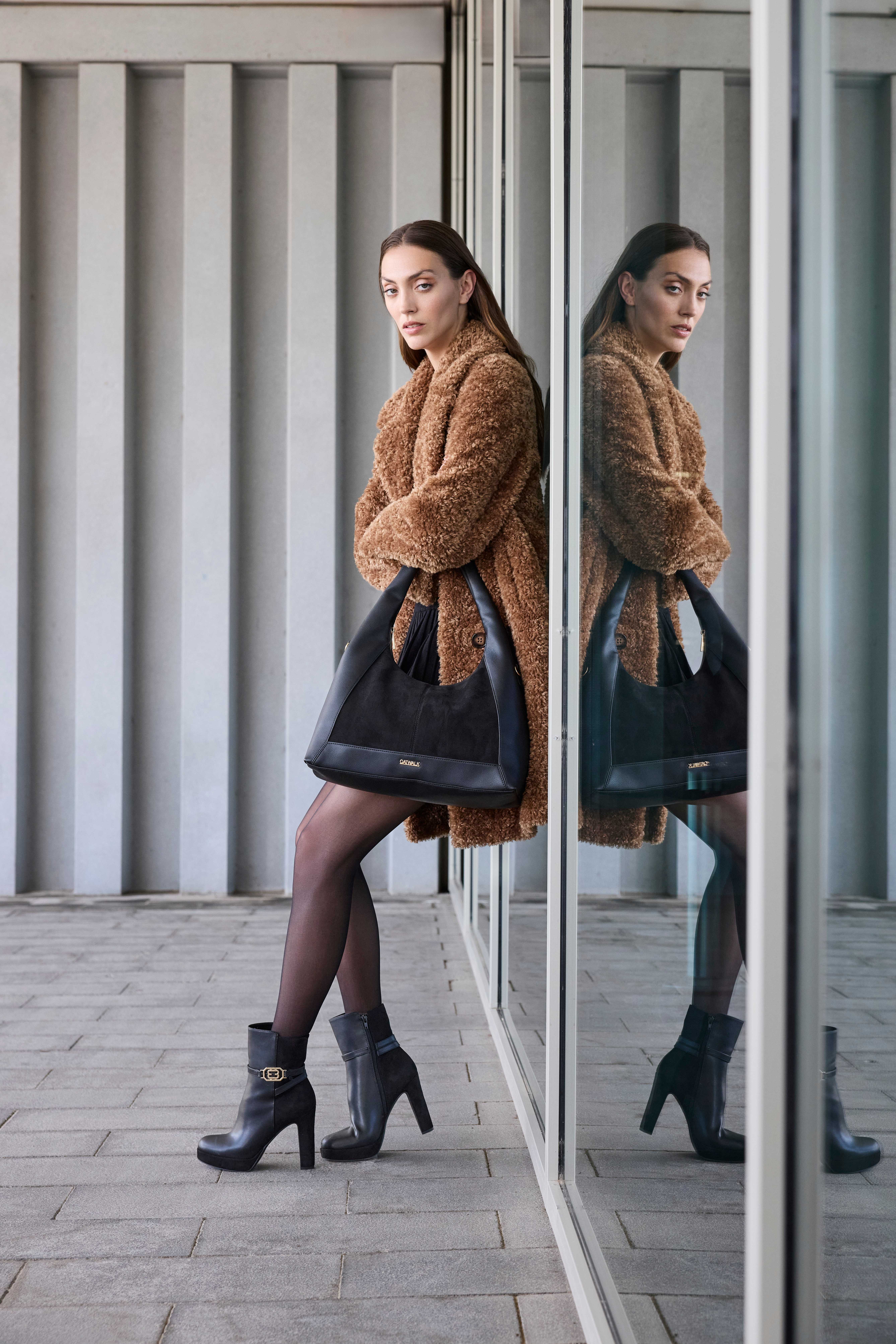 Una donna indossa un abito nero, abbinato a un cappotto marrone in stile teddy, stivaletti neri con tacco e una grande borsa nera
