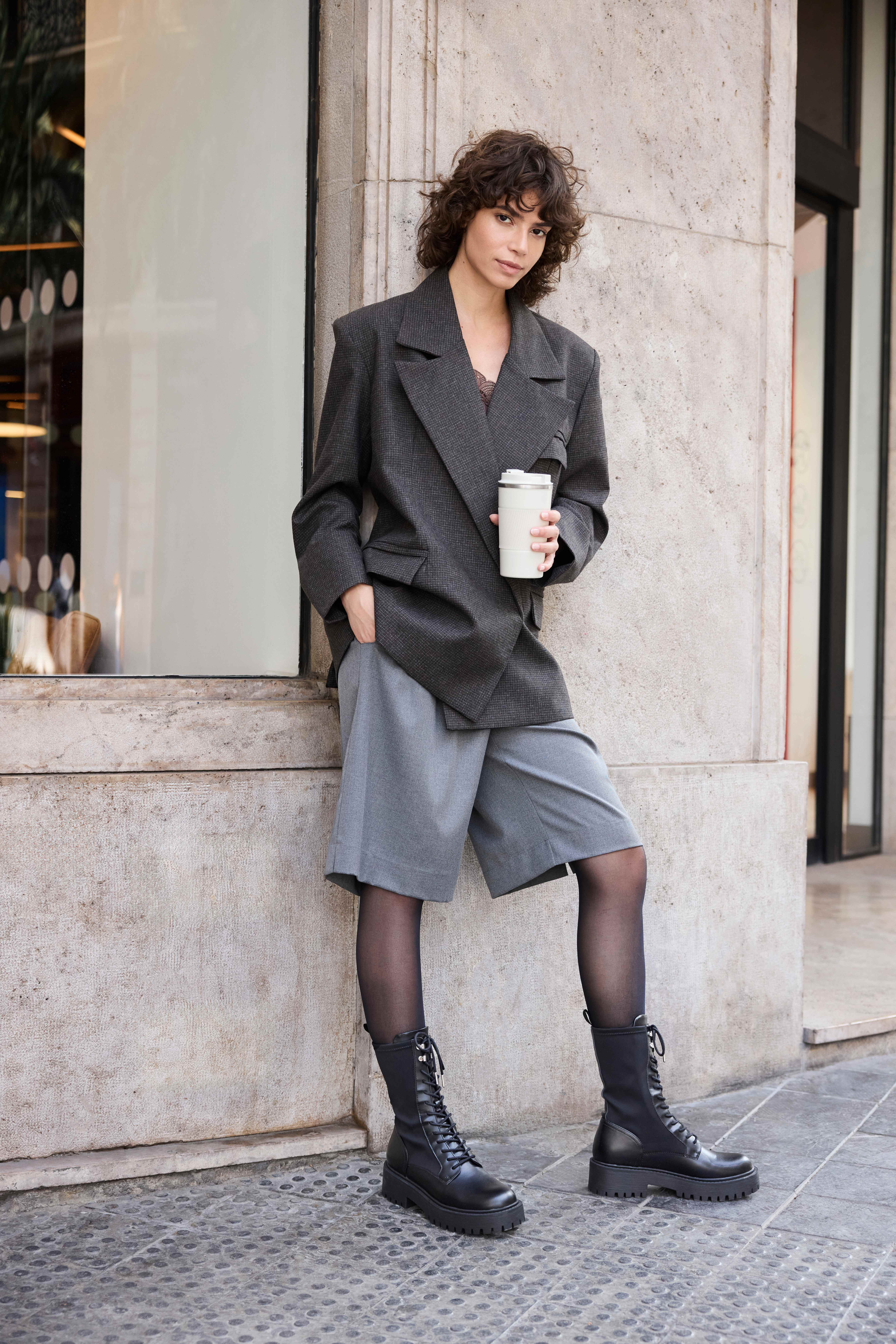 Eine junge Fruau mit braunen Locken, trägt eine schwarze Strumpfhose, einen grauen Blazer, eine graue Shorts und schwarze Schuhe