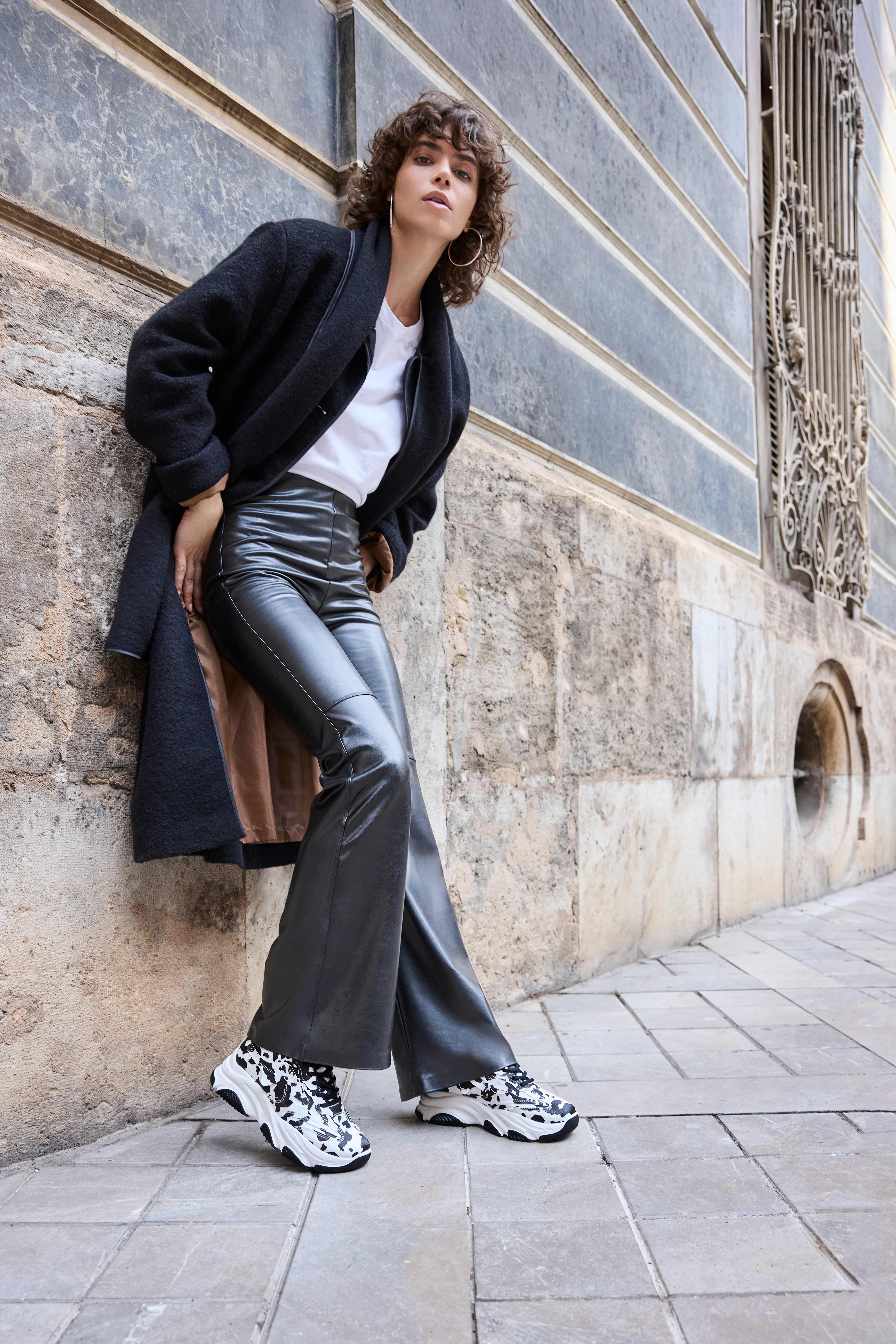 Eine Frau trägt eine schwarze Kunstlederhose mit weitem Schlag, ein weißes T-Shirt, einen langen Mantel und auffällige Sneakers im Kuhfleckenmuster