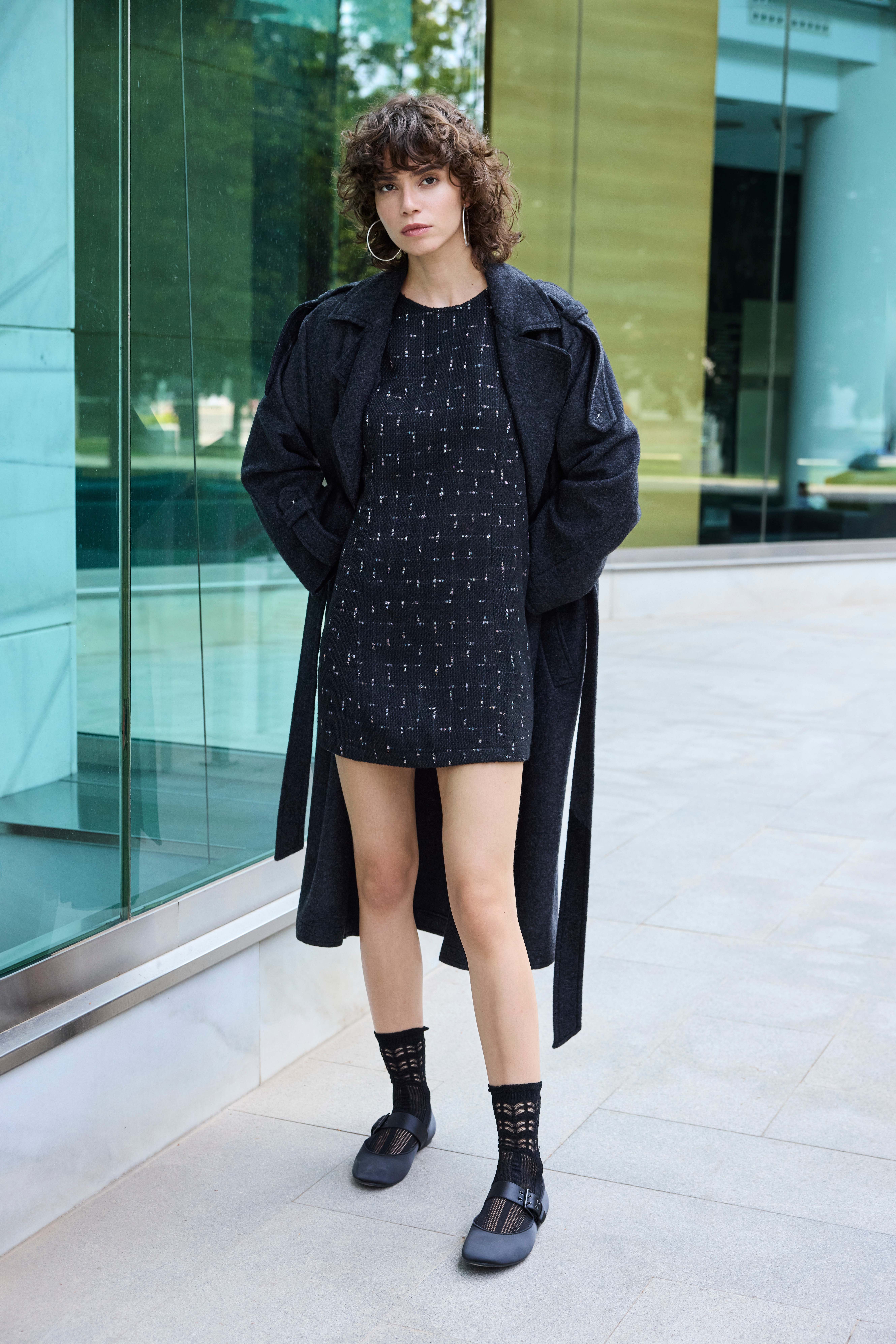 Una donna indossa un miniabito nero a fantasia, un lungo cappotto grigio scuro e ballerine blu scuro con i calzini 